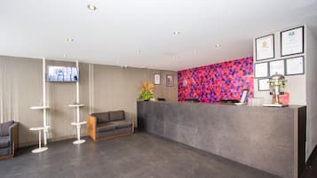 A modern reception area with a grey reception desk, a flat-screen TV mounted on the wall, and a colorful geometric-patterned wall behind the desk.