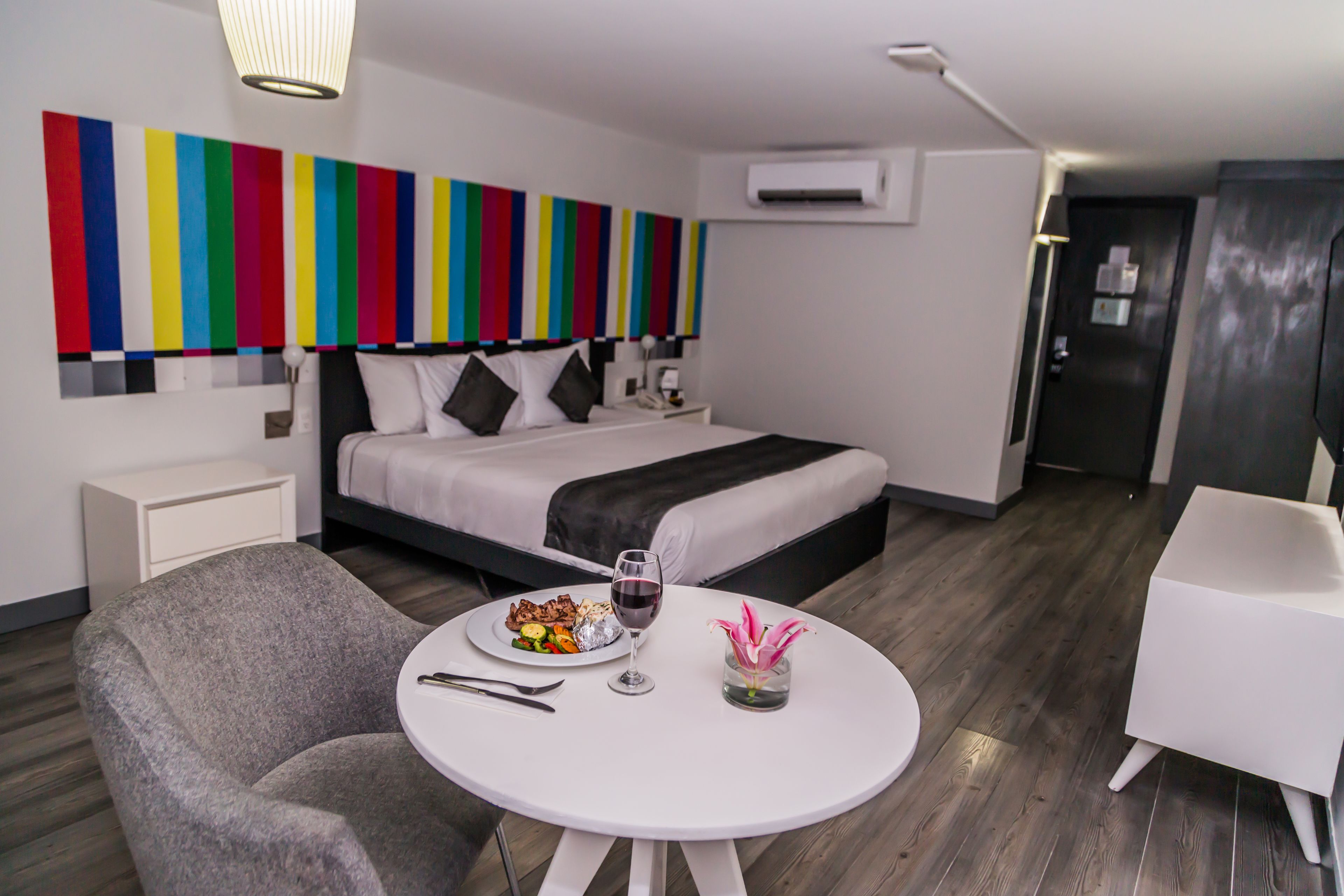 A modern hotel room with a neatly made bed, a dining table with a plate of food and a glass of wine, a gray armchair, and a white round table.