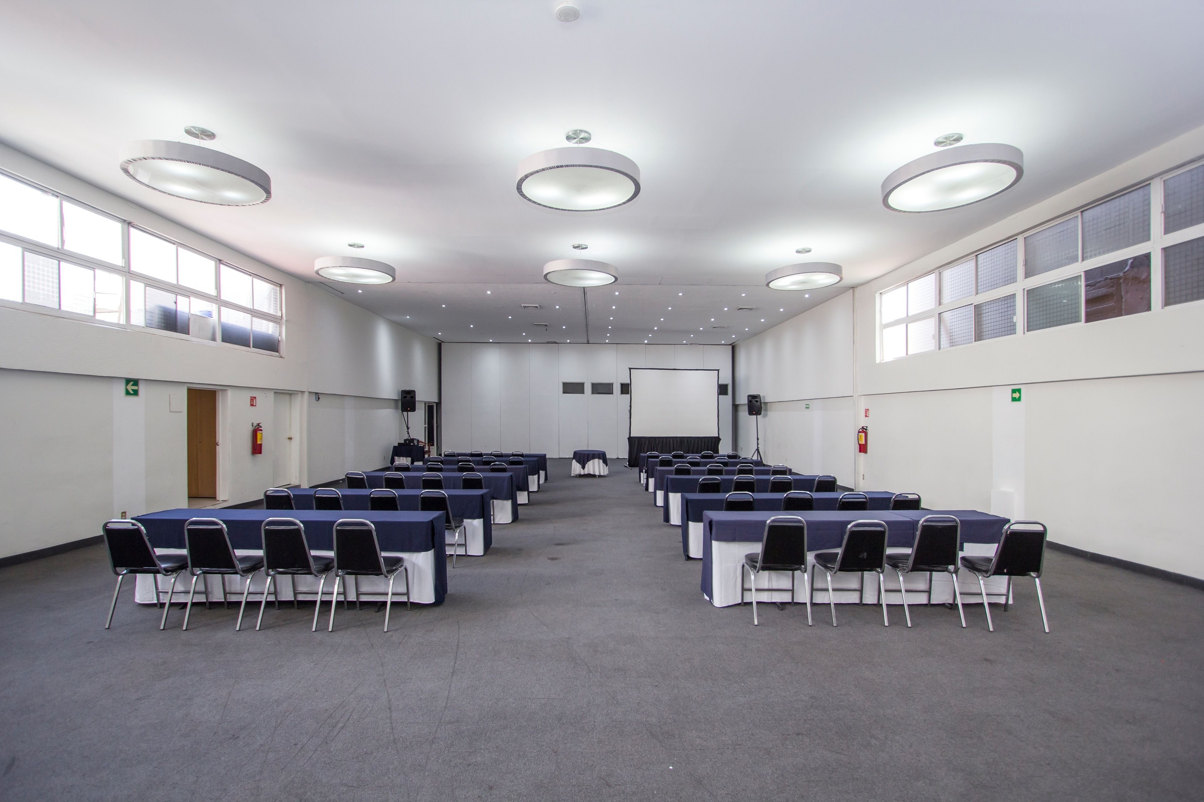 Un salón de conferencias con varias mesas y sillas, ventanas amplias y luces en el techo.