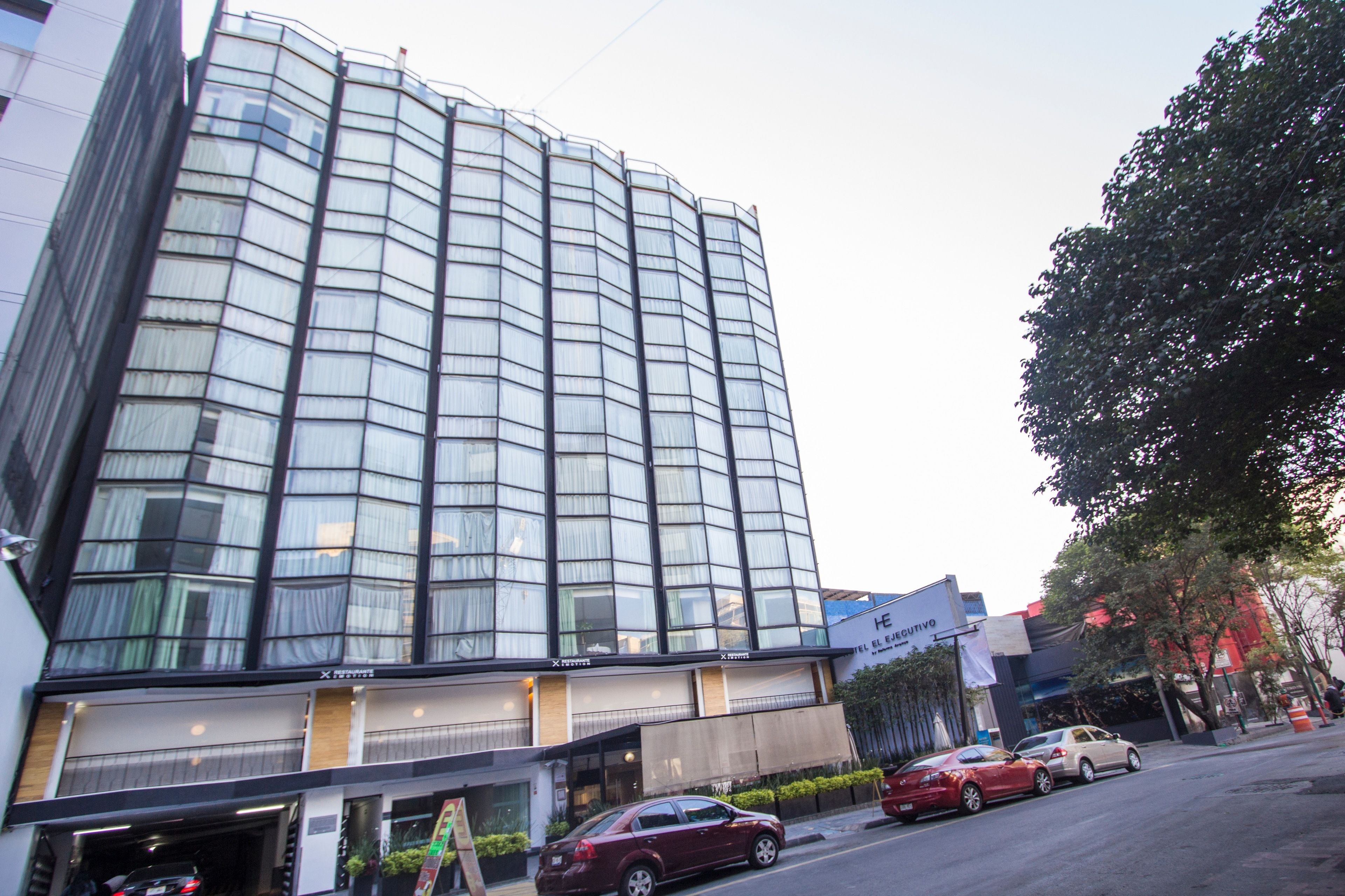 Un hotel moderno con fachada de vidrio curva, varios balcones y vista a la calle con coches estacionados.