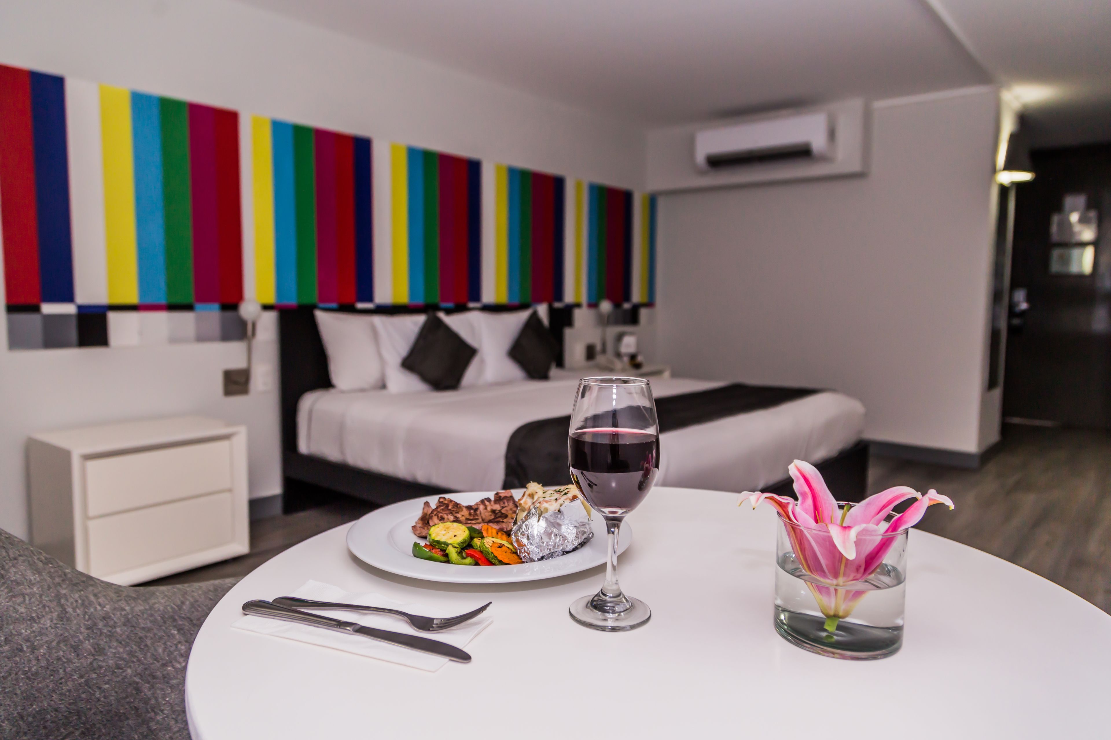 Una habitación de hotel moderna con una cama, una mesa de comedor con un plato de comida y una copa de vino, y una obra de arte de colores en la pared.