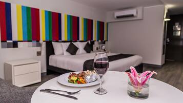 Una habitación de hotel moderna con una cama, una mesa de comedor con un plato de comida y una copa de vino, y una obra de arte de colores en la pared.