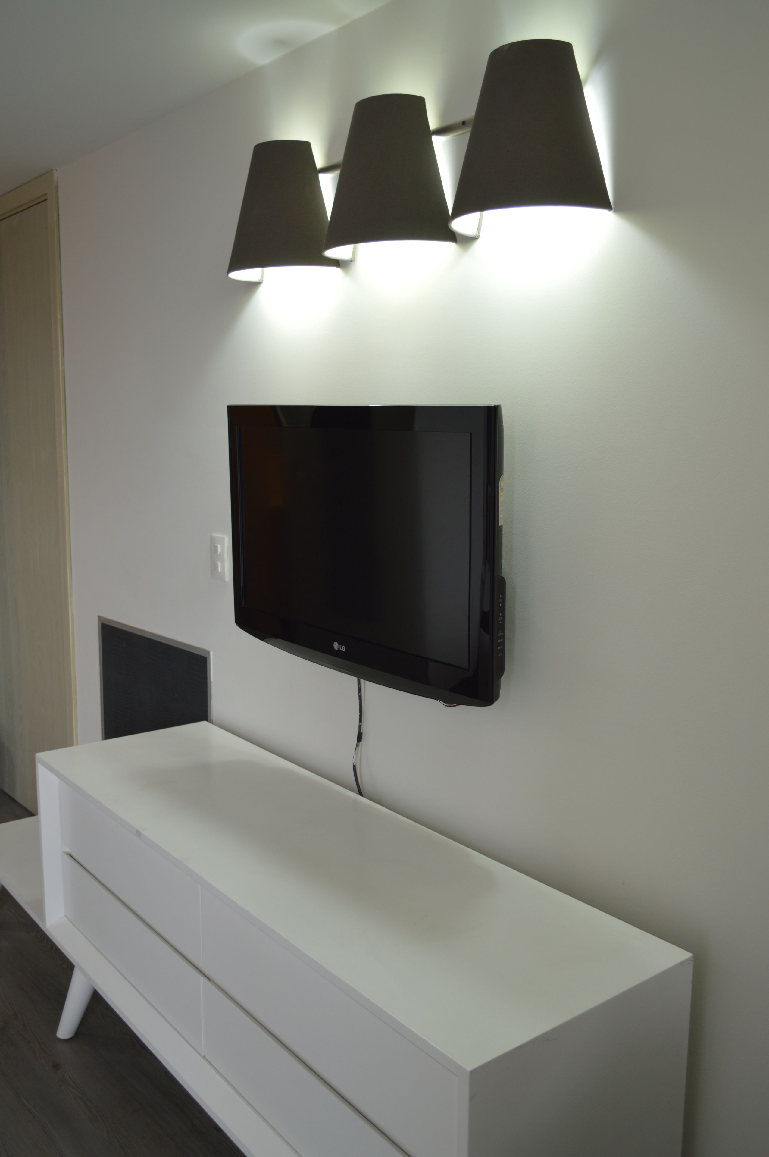 A modern living room with a flat-screen TV mounted on a white wall, flanked by three black wall-mounted lamps.