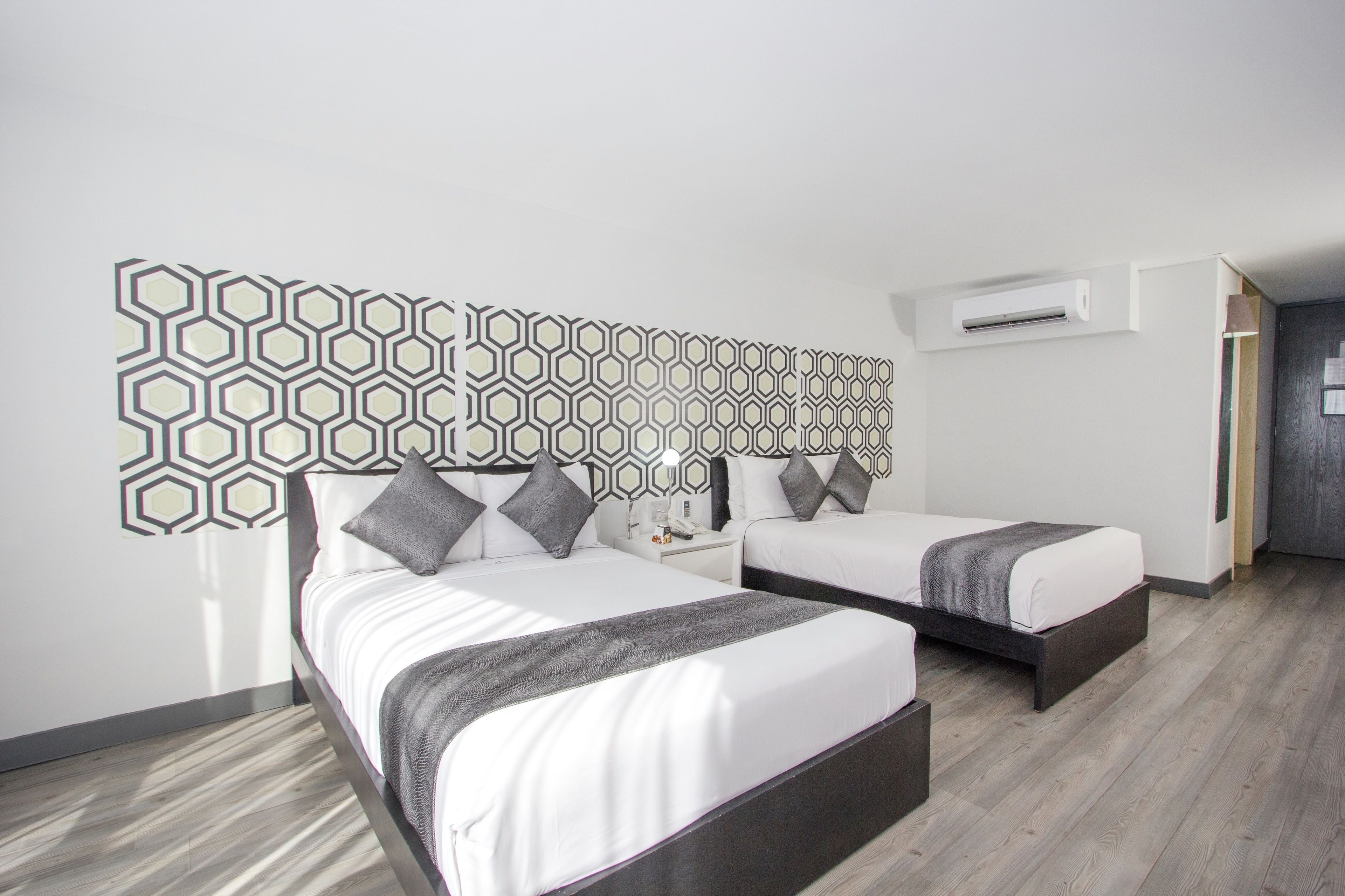 Quarto de hotel moderno com duas camas, design geométrico na parede, piso de madeira e ar condicionado.