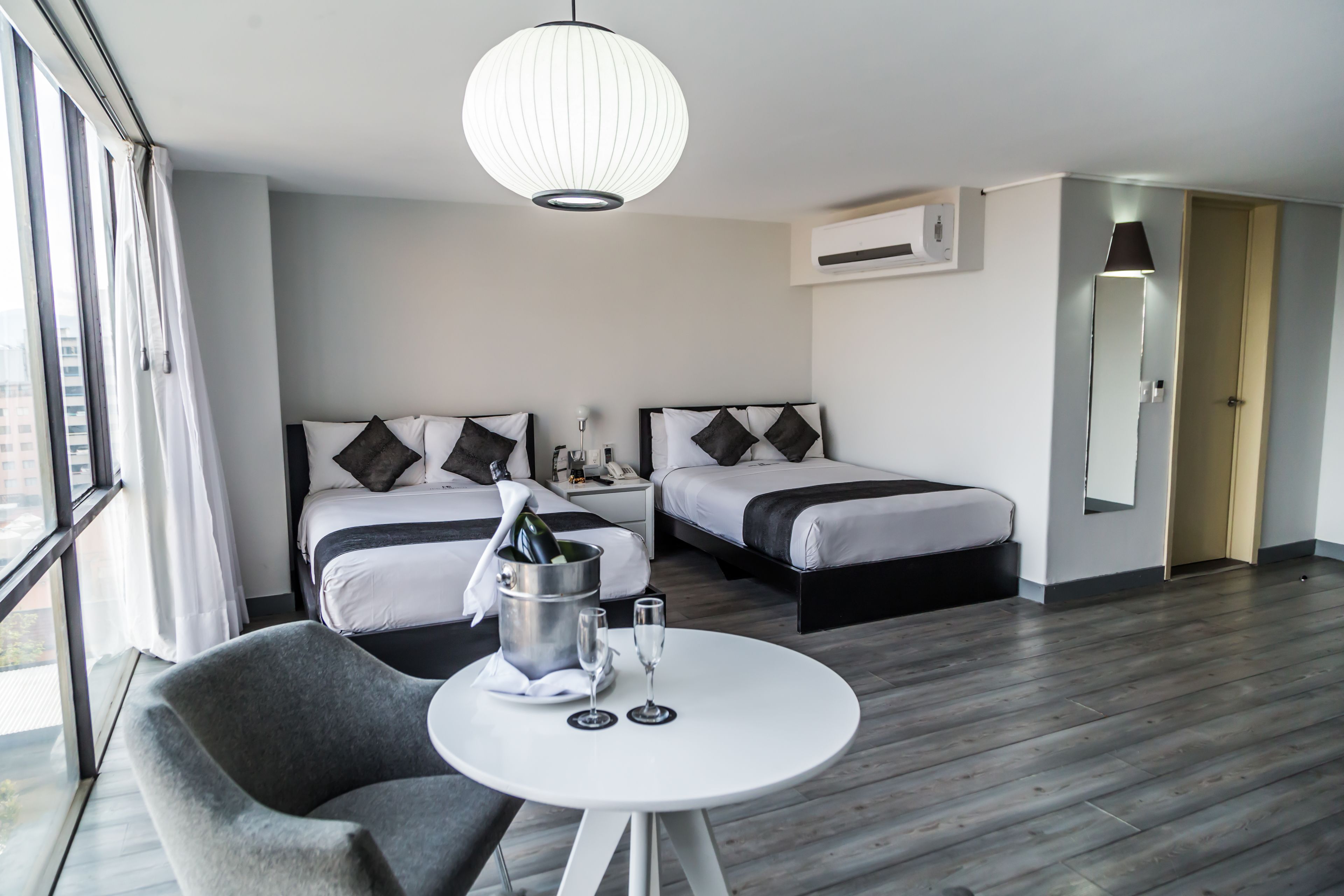 A hotel room with two beds, a round table with a bottle and glasses, a grey armchair, and a large window.