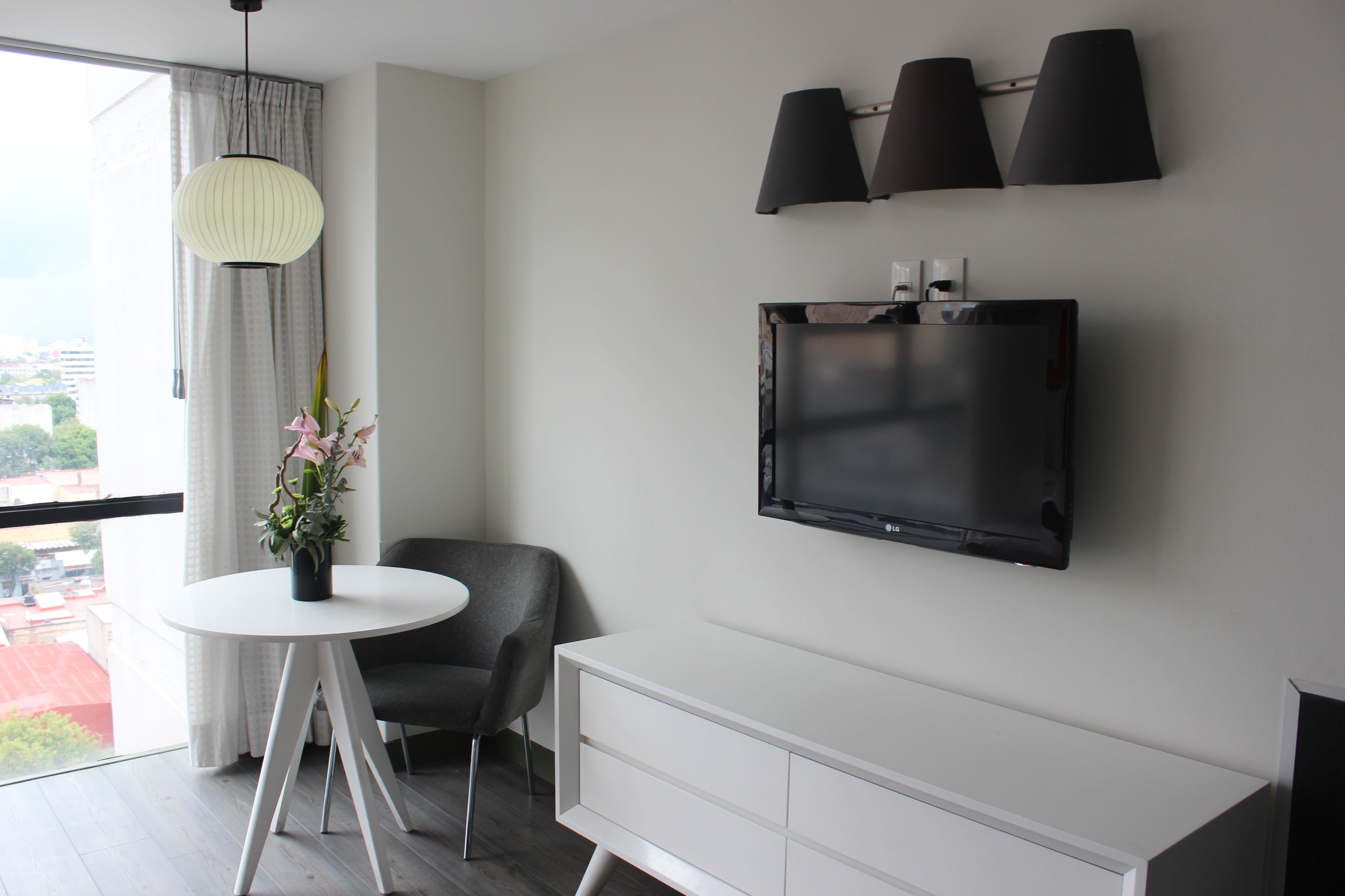 Una habitación moderna con un ventanal grande, una mesa redonda con un florero, una silla gris, un armario blanco y un televisor de pared.