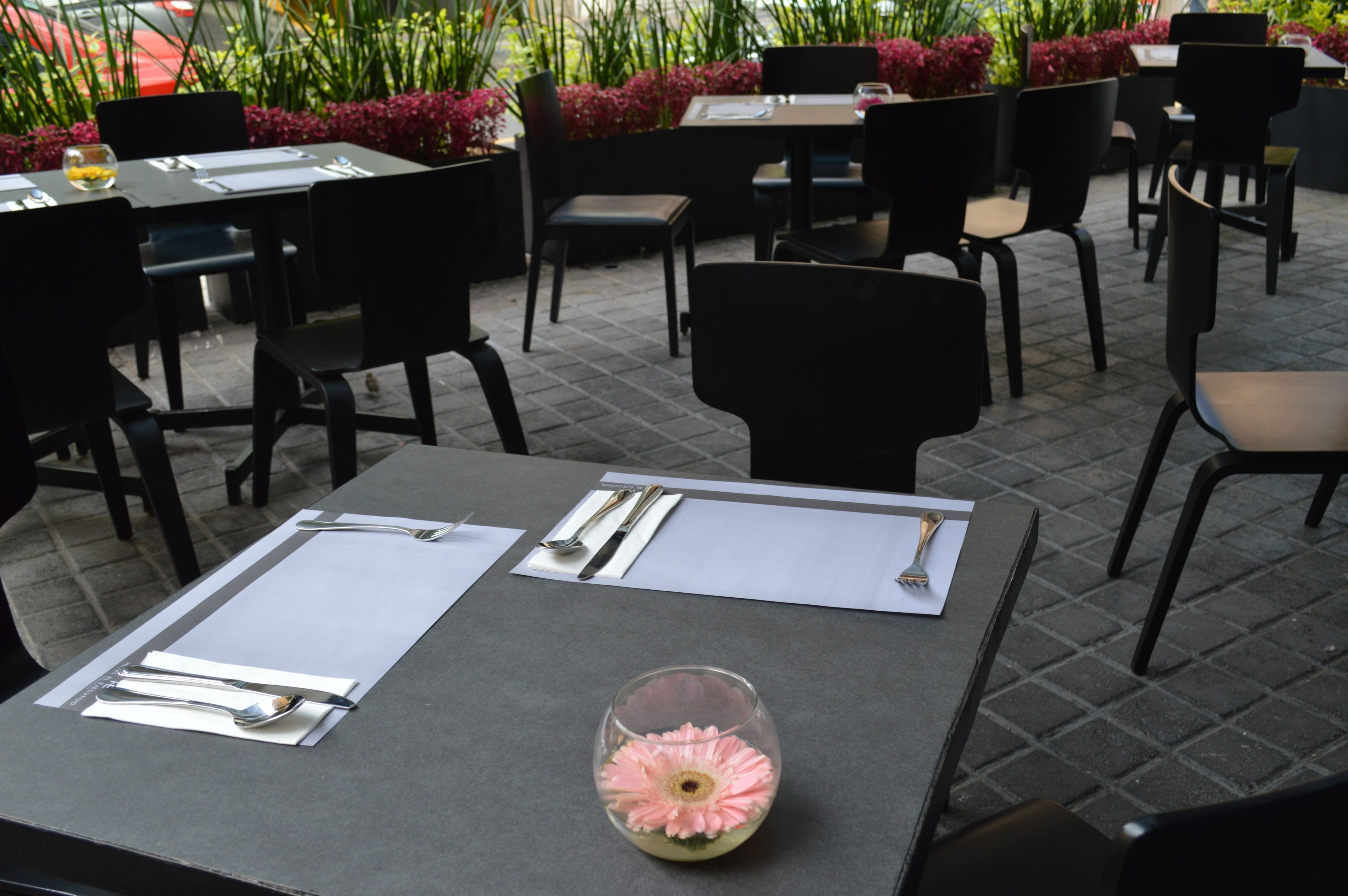 Un área de comedor al aire libre con mesas preparadas para dos, sillas negras y un jarrón de cristal con un centro de mesa de flor rosa.
