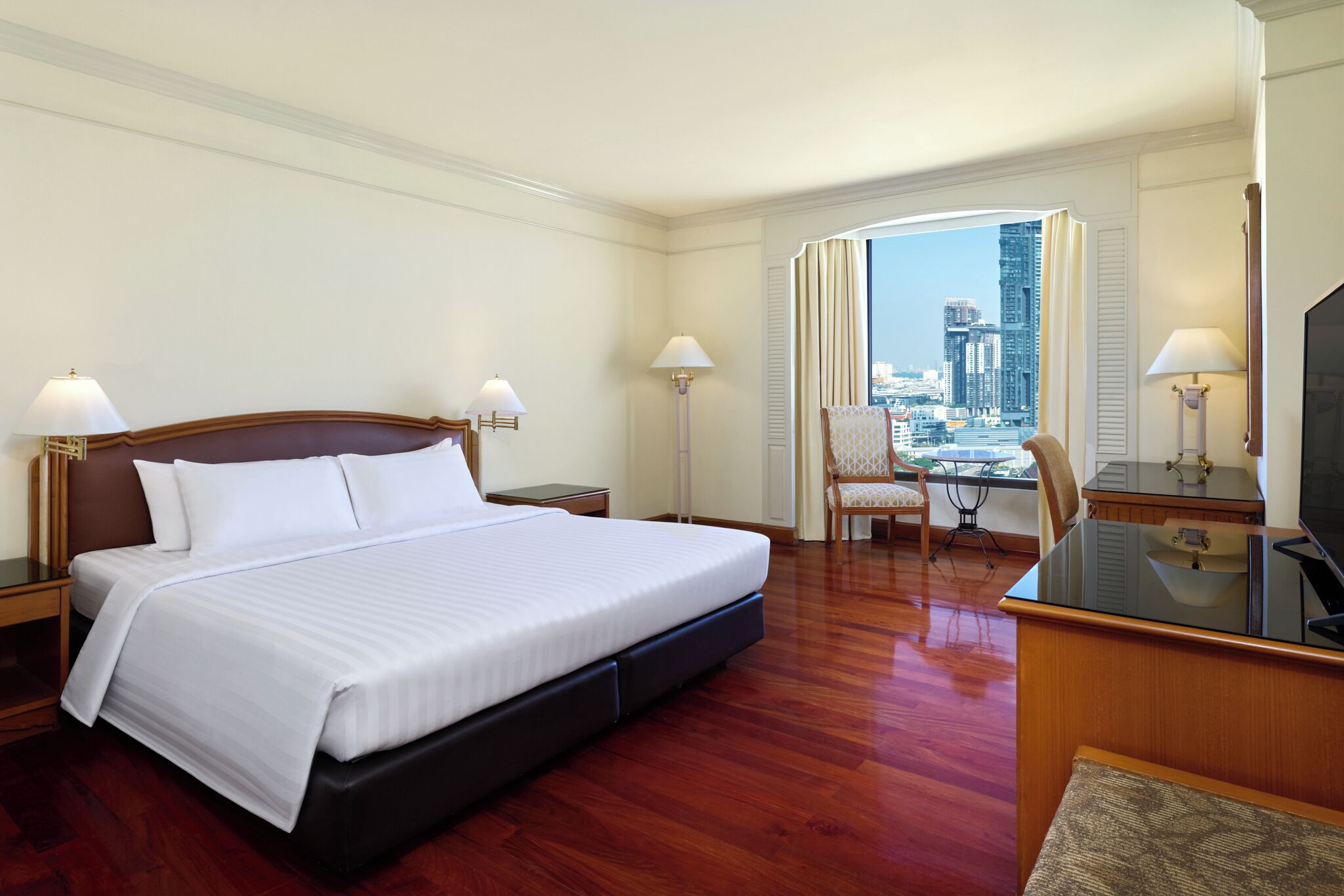 A hotel room with a bed, bedside tables, a desk with a TV, a chair, and a view of a cityscape.