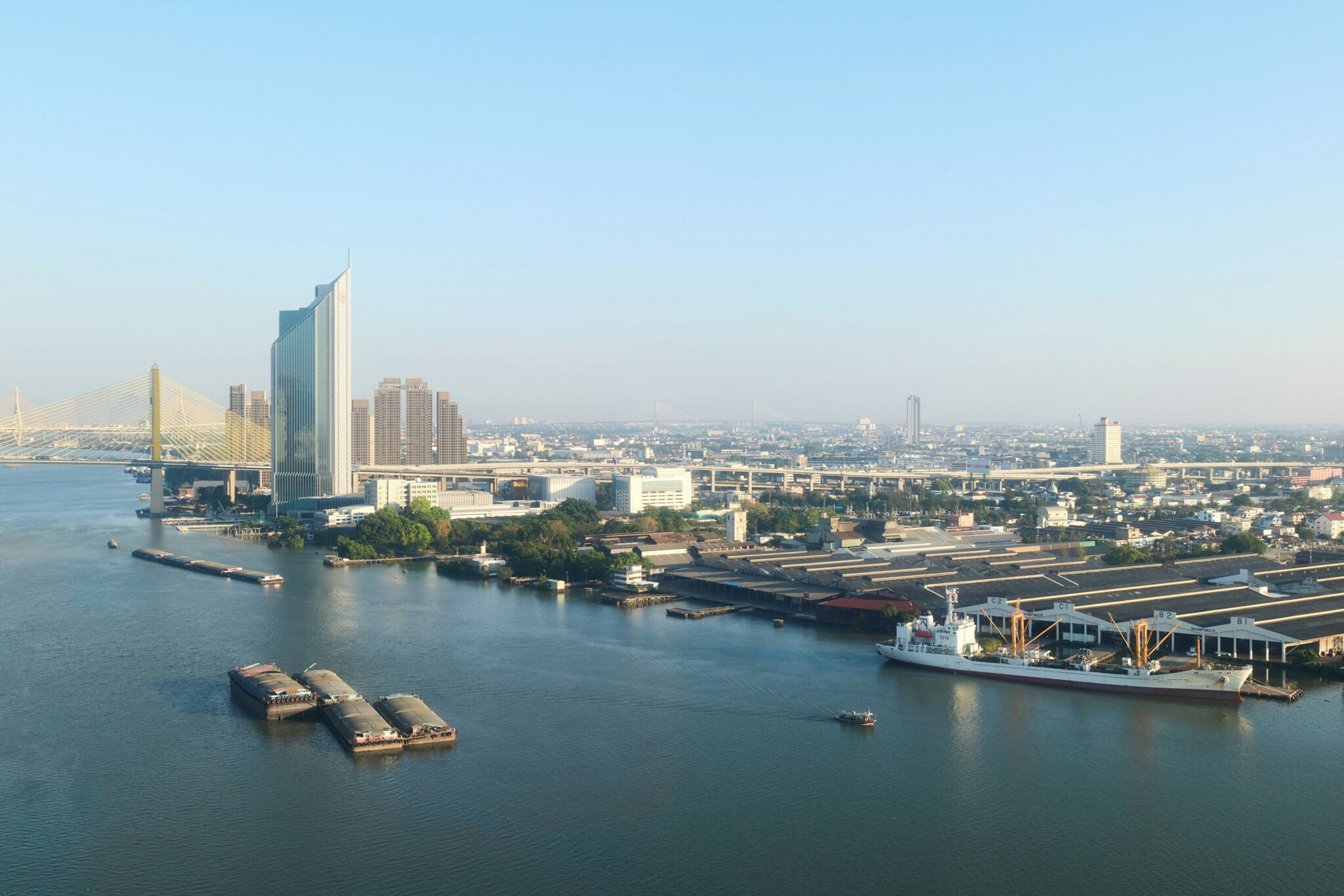 A riverfront cityscape with a prominent bridge, high-rise buildings, and a large cargo ship docked at a port.
