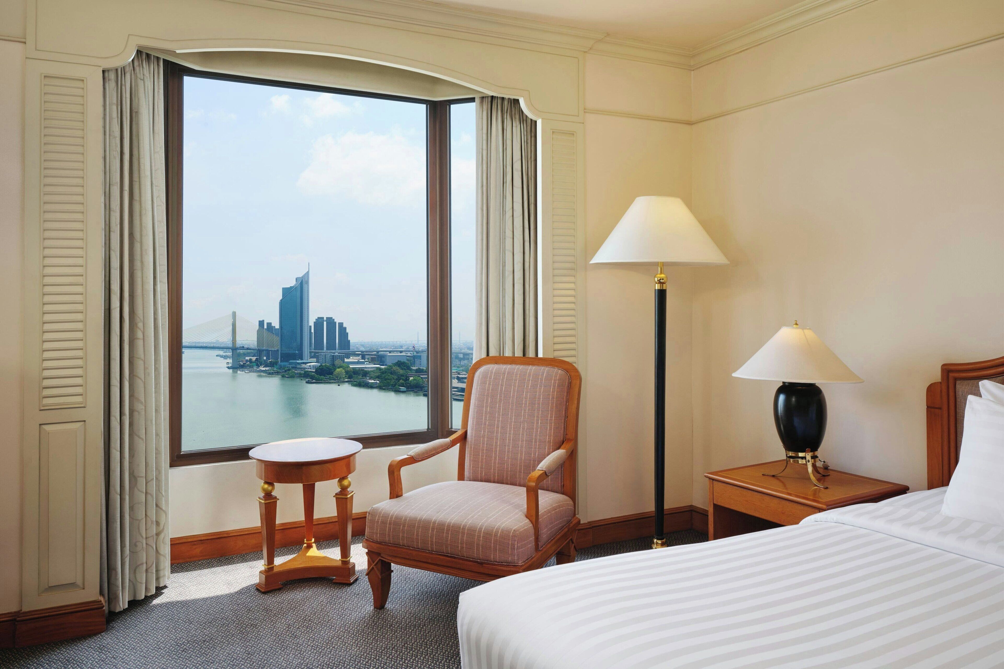 A hotel room with a large window, a bed, a chair, a nightstand, and a lamp.