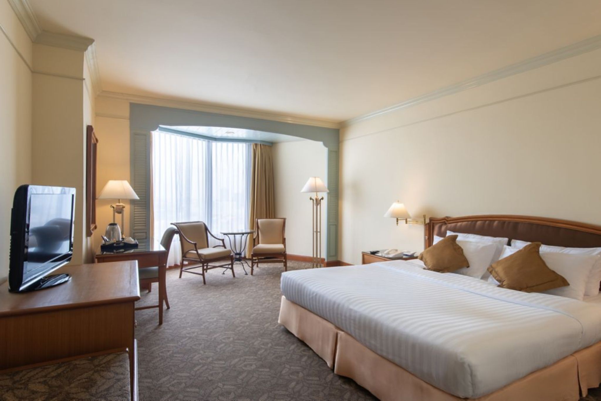 A hotel room with a bed, a desk with a television, two chairs, a small table, lamps, and a window with curtains.