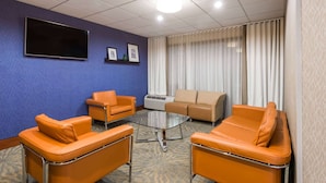 A room with a blue wall, a flat-screen TV mounted above a glass table surrounded by orange chairs, a beige sofa, and a large window with white curtains.