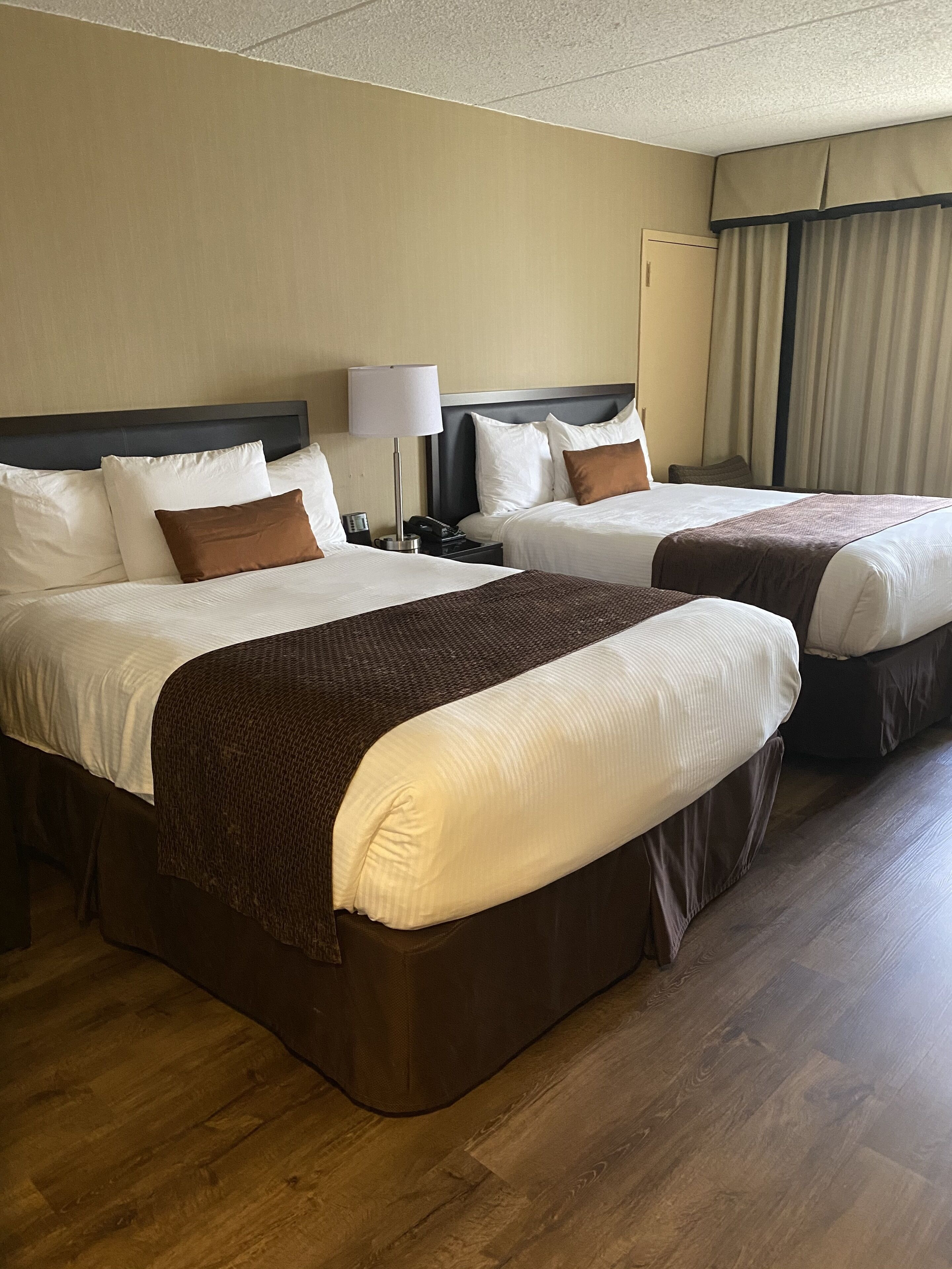 A hotel room with two beds, a bedside table with a lamp, and a window with curtains.
