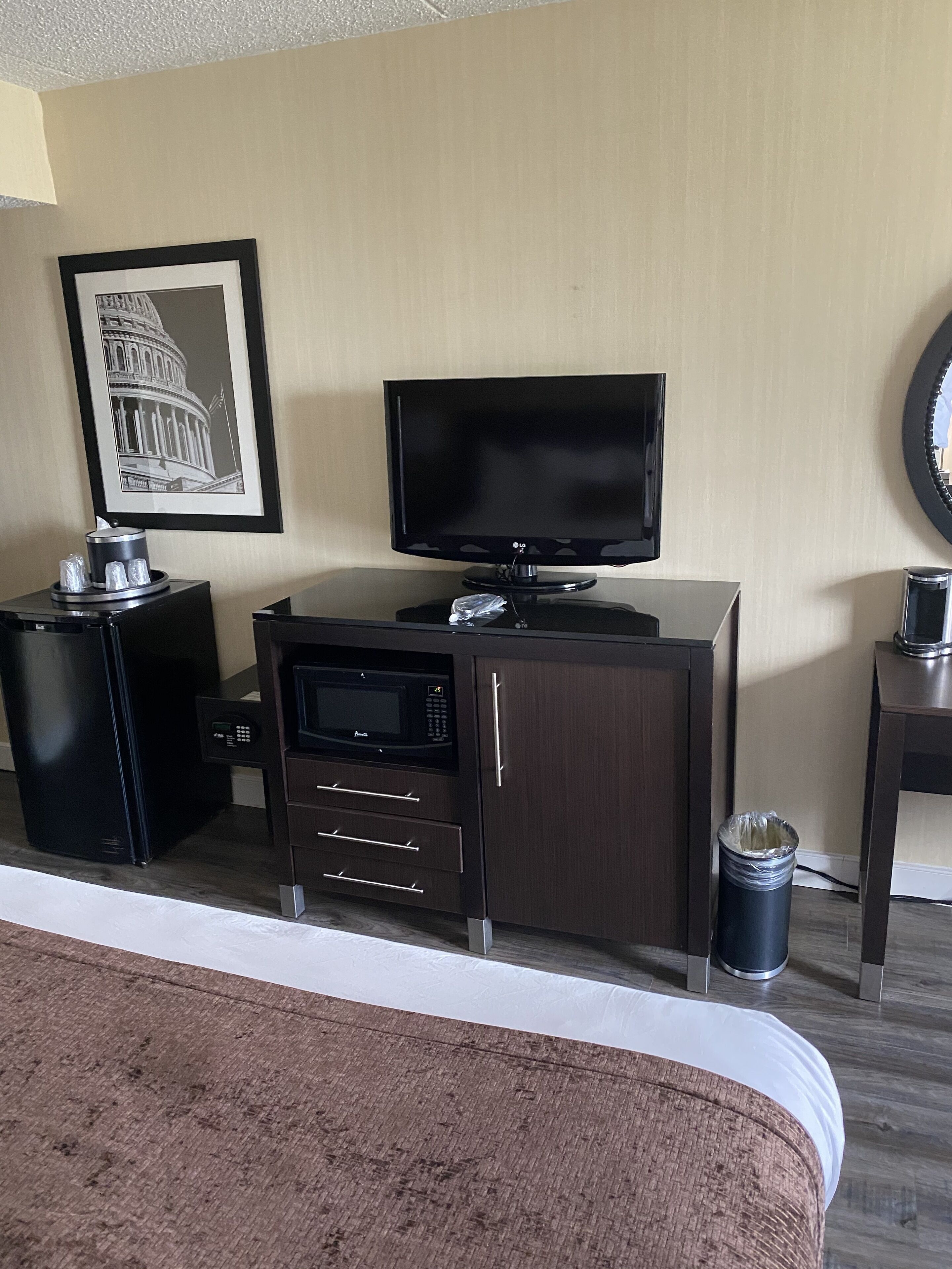 A hotel room with a bed, a television, a microwave, a small refrigerator, a trash can, and a framed picture of the Capitol building.