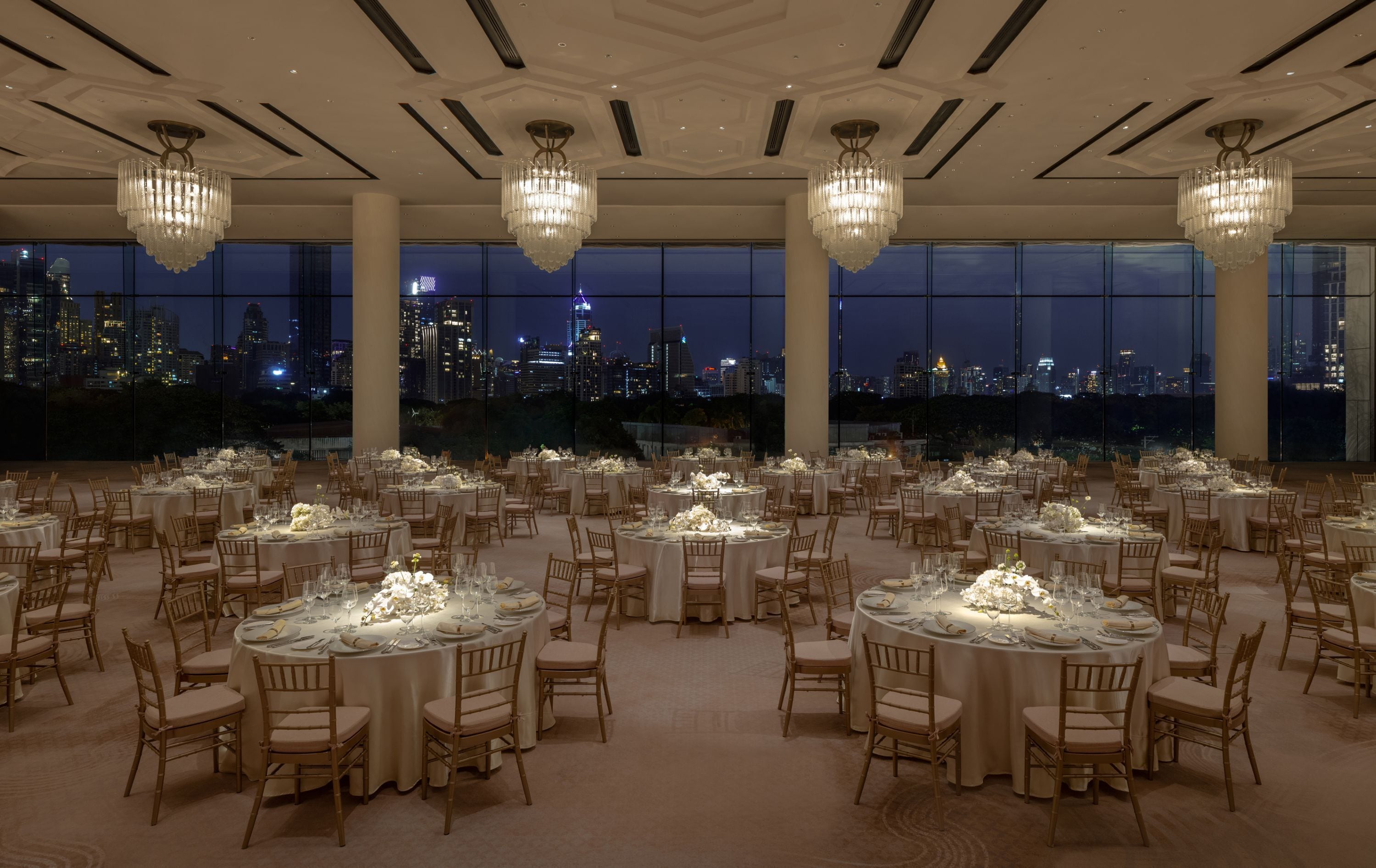 A banquet hall with round tables set for a formal event, large windows offering a cityscape view, and ornate chandeliers.