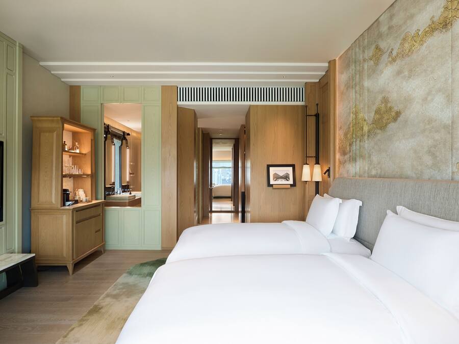A hotel room with a large bed, a TV, a desk with a mirror, and a view of the city through the window.