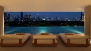 A modern hotel room with a city skyline view, an infinity pool, and three white sofas.