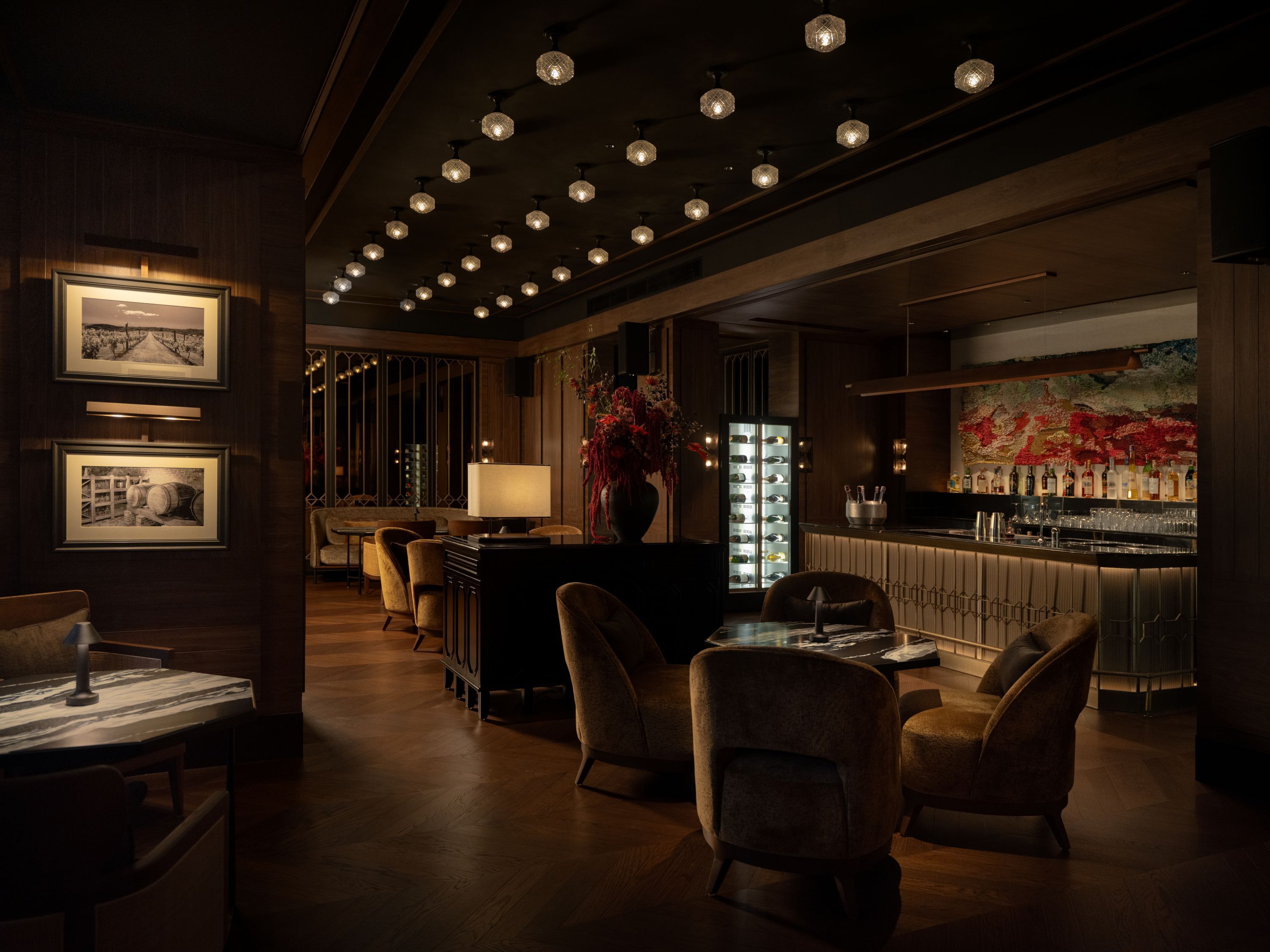 A dimly lit bar area with wooden flooring, bar stools, and a large wine refrigerator.