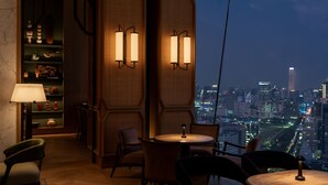 A dimly lit room with a city view, featuring a round table, chairs, a lamp, and wall-mounted lights. - Dusit Thani Bangkok (Bangkok)
