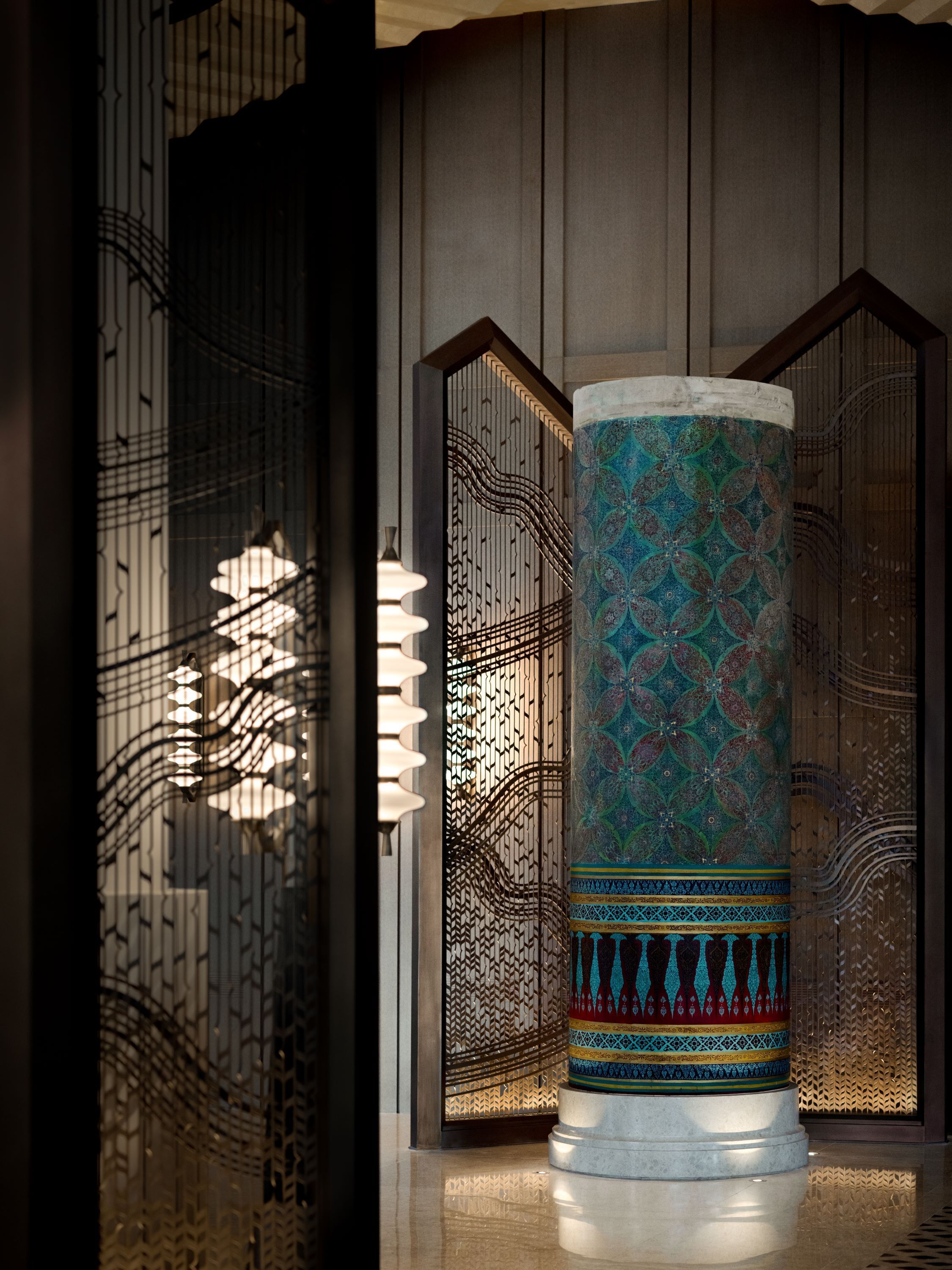 Une colonne décorative ornée de motifs complexes et dotée d’une base réfléchissante, placée devant un décor de murs à motifs et un treillis.