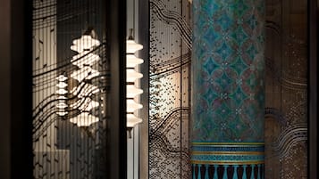 Une colonne décorative ornée de motifs complexes et dotée d’une base réfléchissante, placée devant un décor de murs à motifs et un treillis.