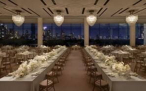 A banquet hall with tables set for a formal event, large windows offering a city view, and elegant chandeliers.
