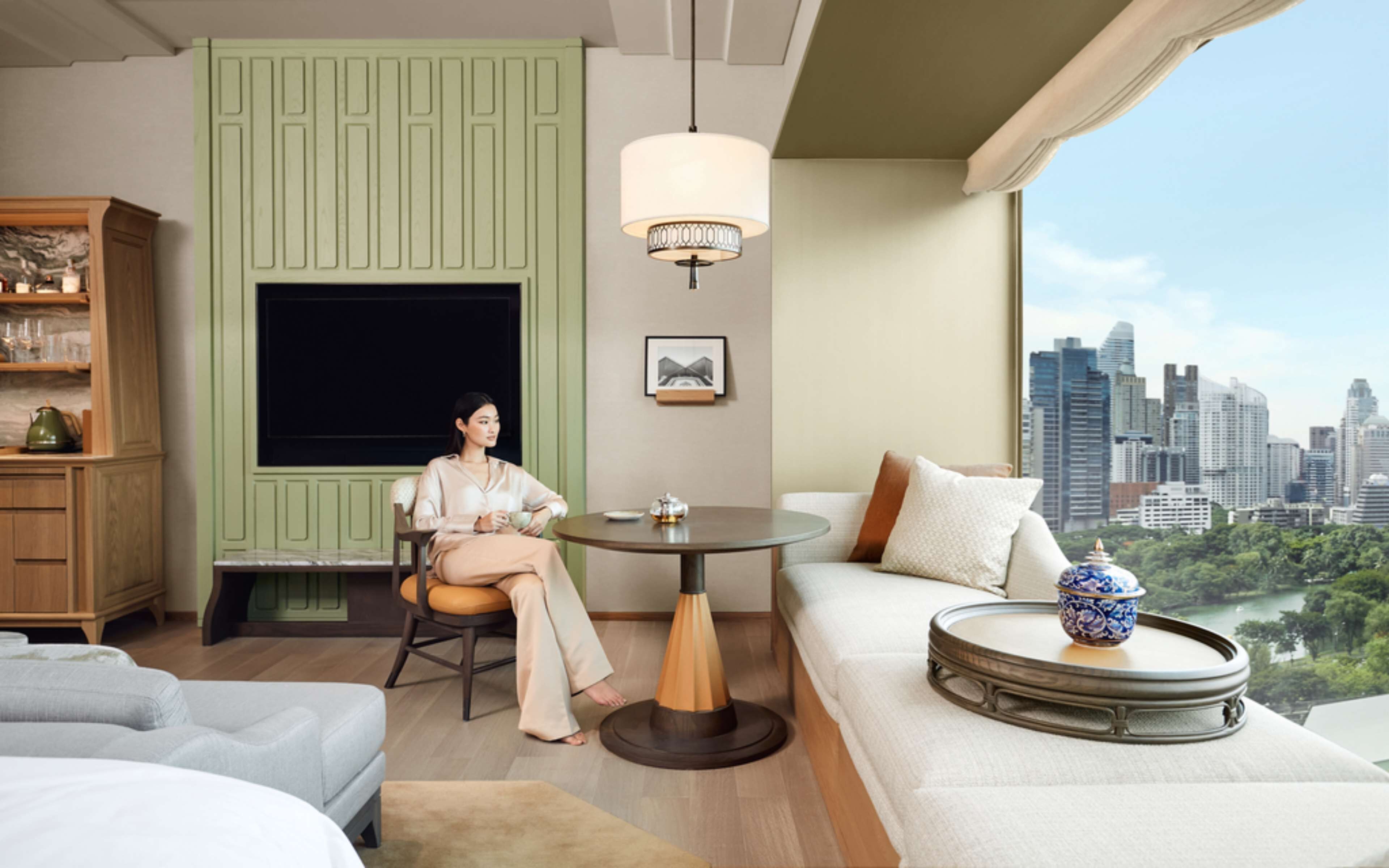 A modern hotel room with a city view, a green accent wall, a round table, and a sofa with a decorative bowl.