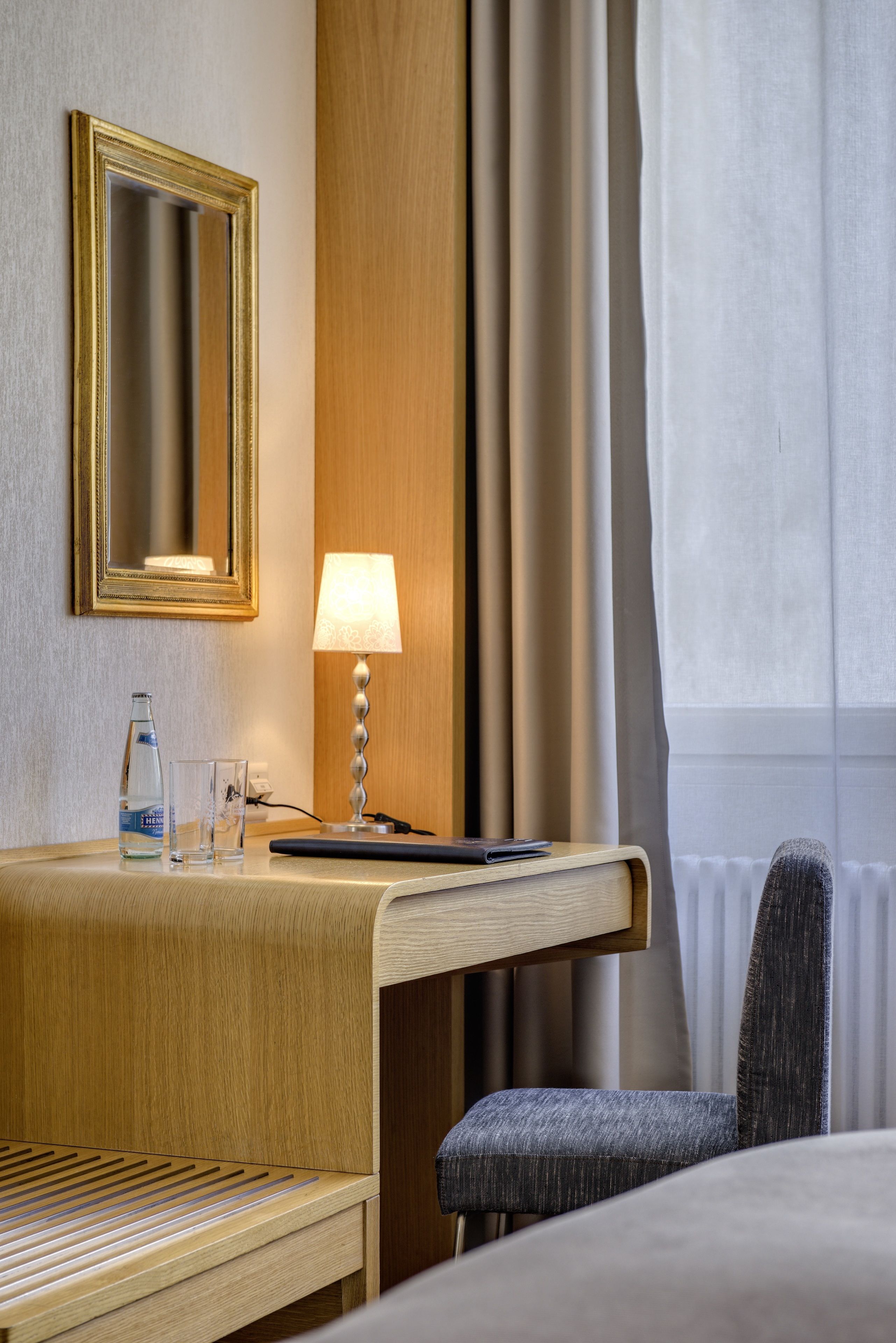 A hotel room with a wooden desk, a lamp, a bottle of water, and a chair.