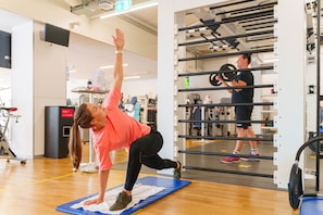 Un gimnasio con equipo de ejercicio, una persona realizando una postura de yoga sobre una esterilla y otra persona levantando pesas.