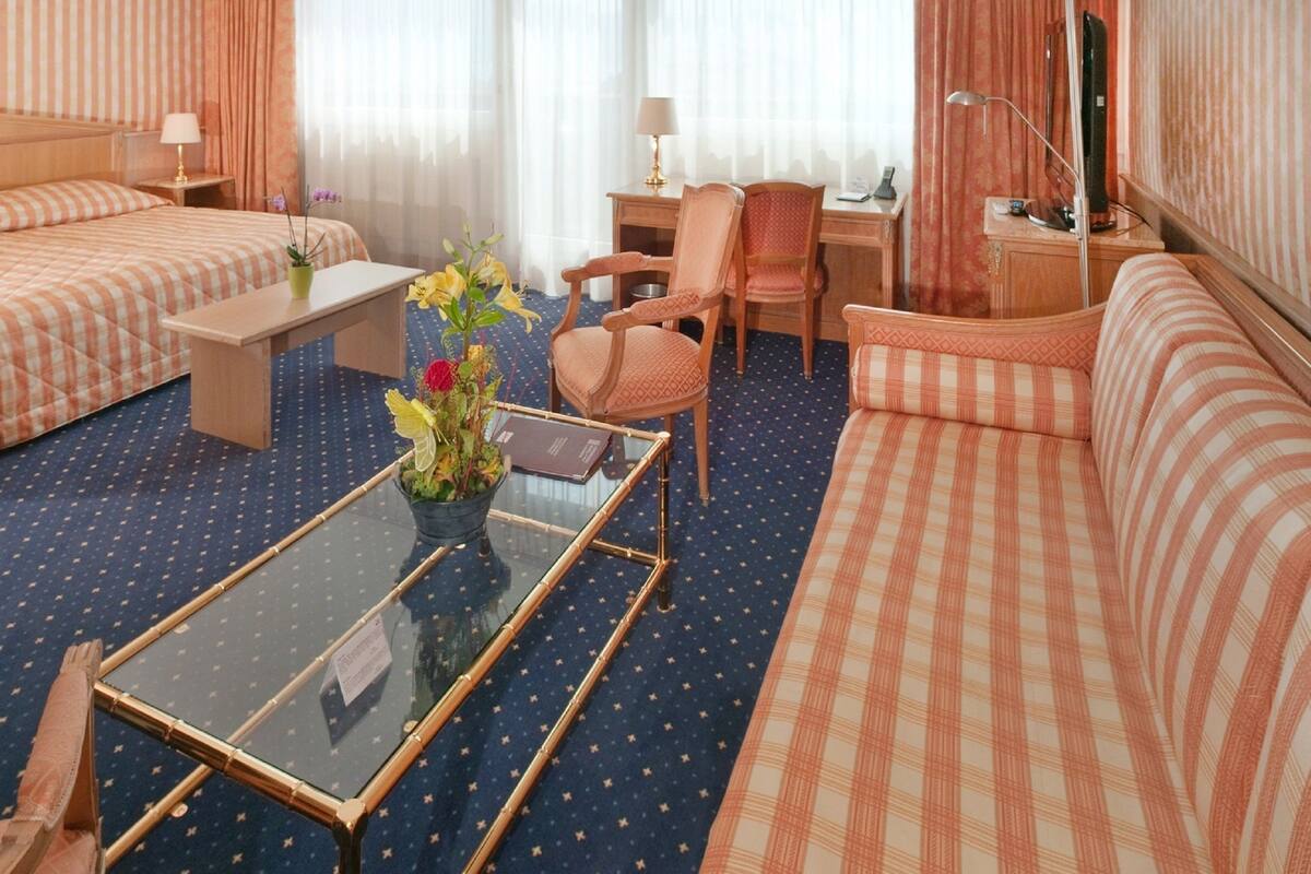 Hotellihuone, jossa on sänky, sohva, pieni pöytä maljakko täynnä kukkia, tuoli, työpöytä lampun kanssa ja televisio.