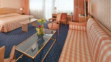 A hotel room with a bed, a sofa, a small table with a vase of flowers, a chair, a desk with a lamp, and a television.