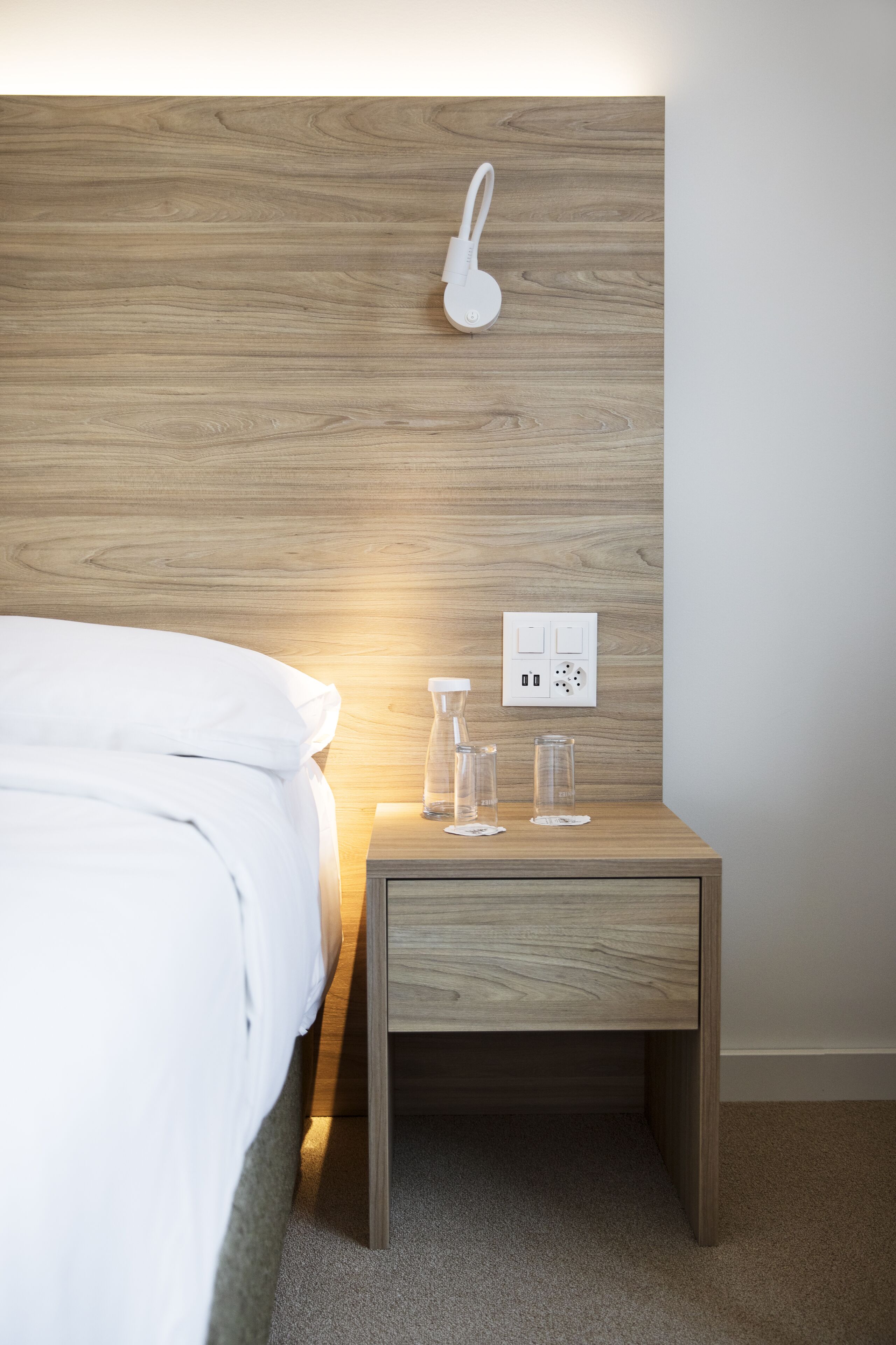 A neatly made bed with white linens, a wooden bedside table with a white lamp, and a power outlet on the wall.