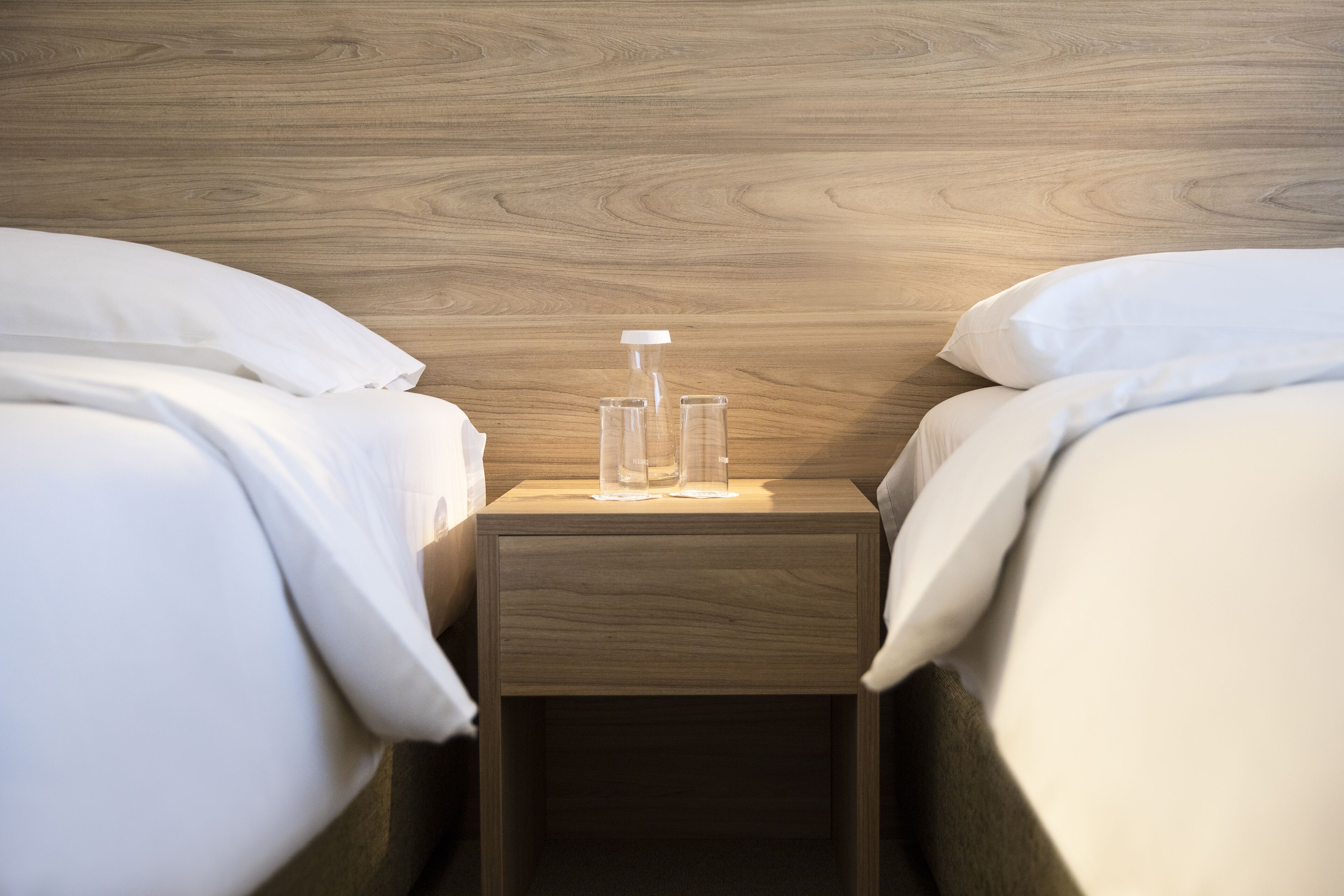 A hotel room with two beds, a wooden headboard, and a nightstand with a glass bottle and a glass.