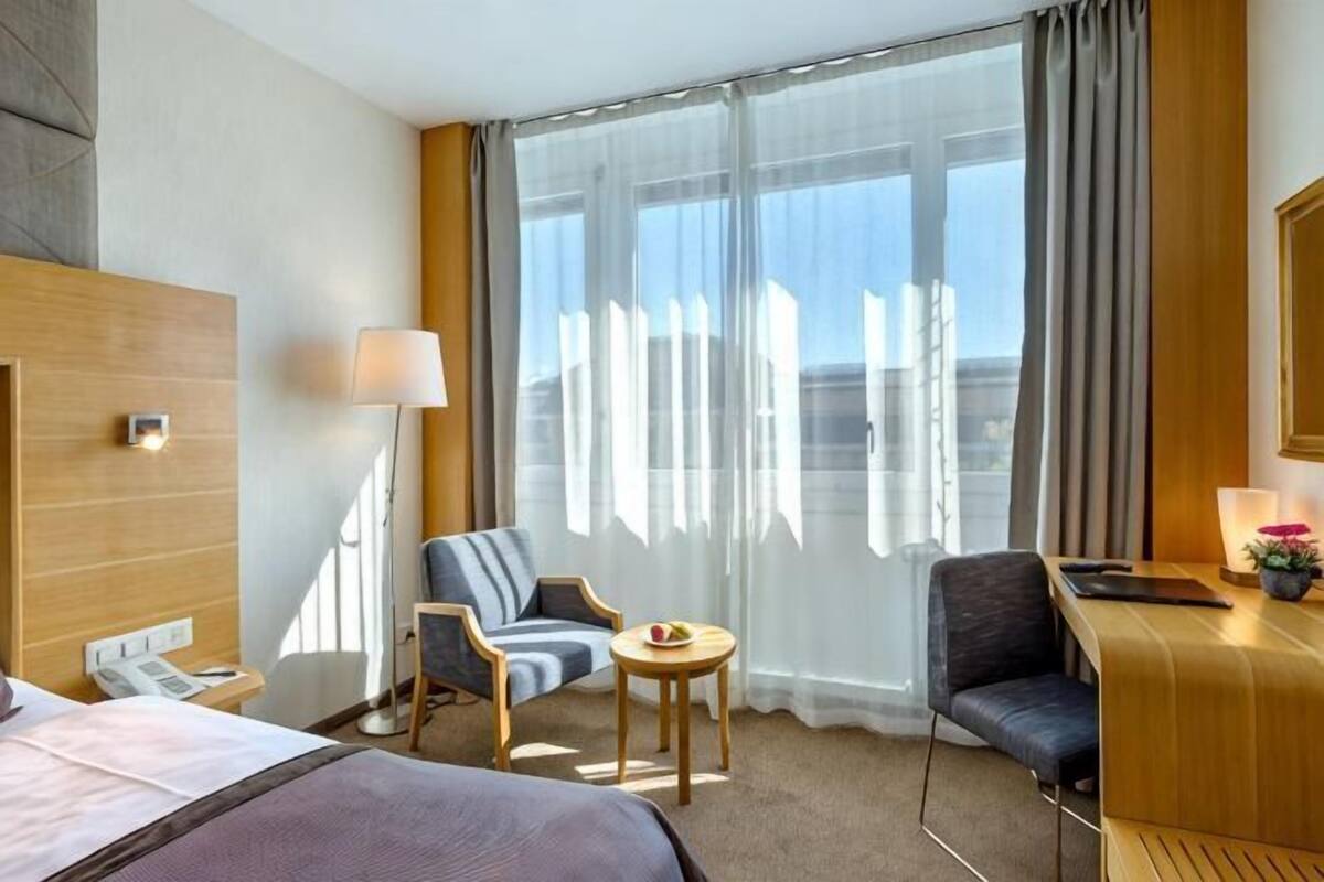 Hotellihuone, jossa on sänky, kaksi nojatuolia, pieni pöytä, työpöytä lampun kanssa ja suuri ikkuna, jossa on verhot.