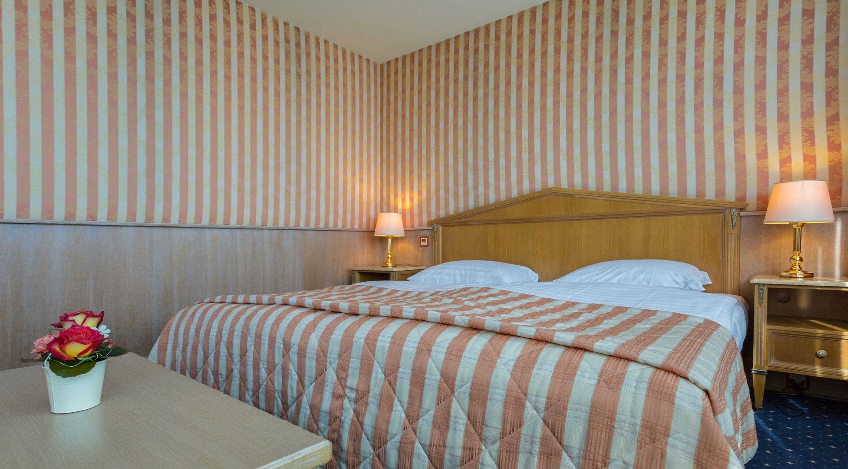Una camera d'albergo con un letto, due comodini, una lampada e un vaso con fiori.