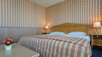 Habitació d'hotel amb un llit, dues taules de nit, una petita taula auxiliar, una làmpada i un vas amb flors.