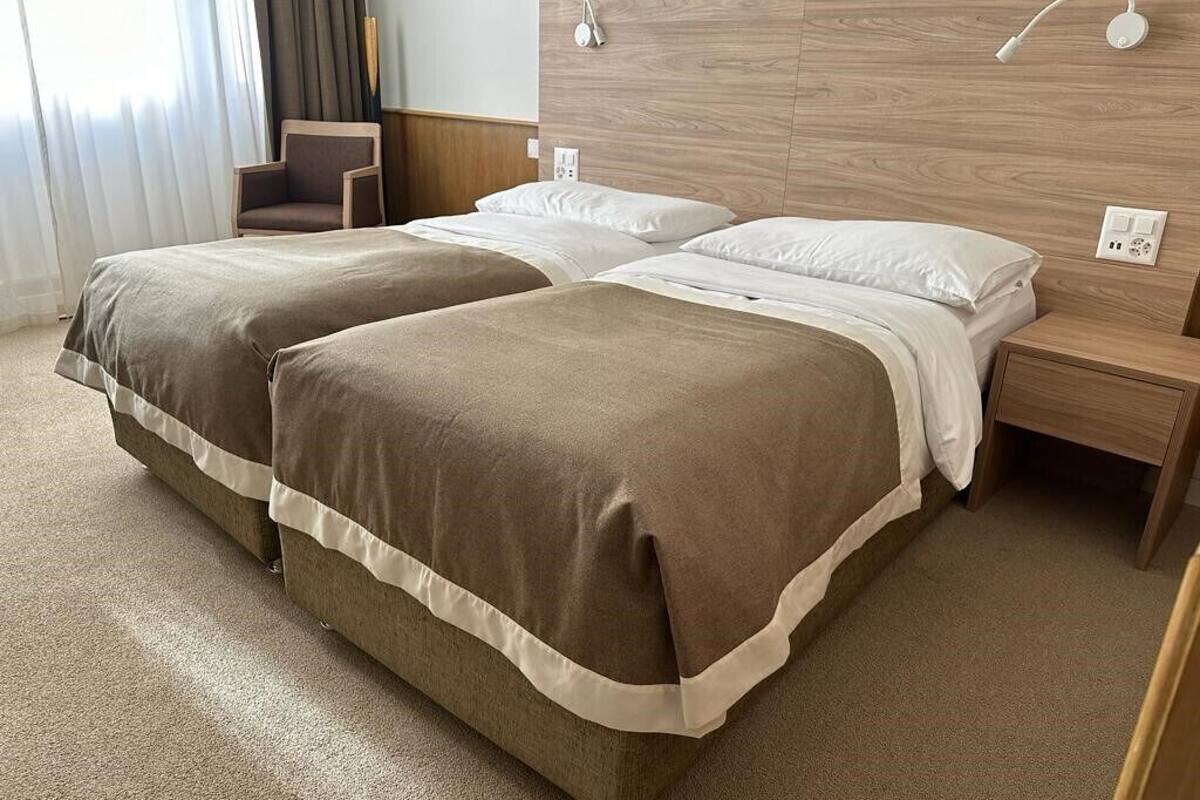 Hotellihuone, jossa on kaksi sänkyä, puinen pääty, yöpöytävalaisimet, tuoli ja matolla peitetty lattia.