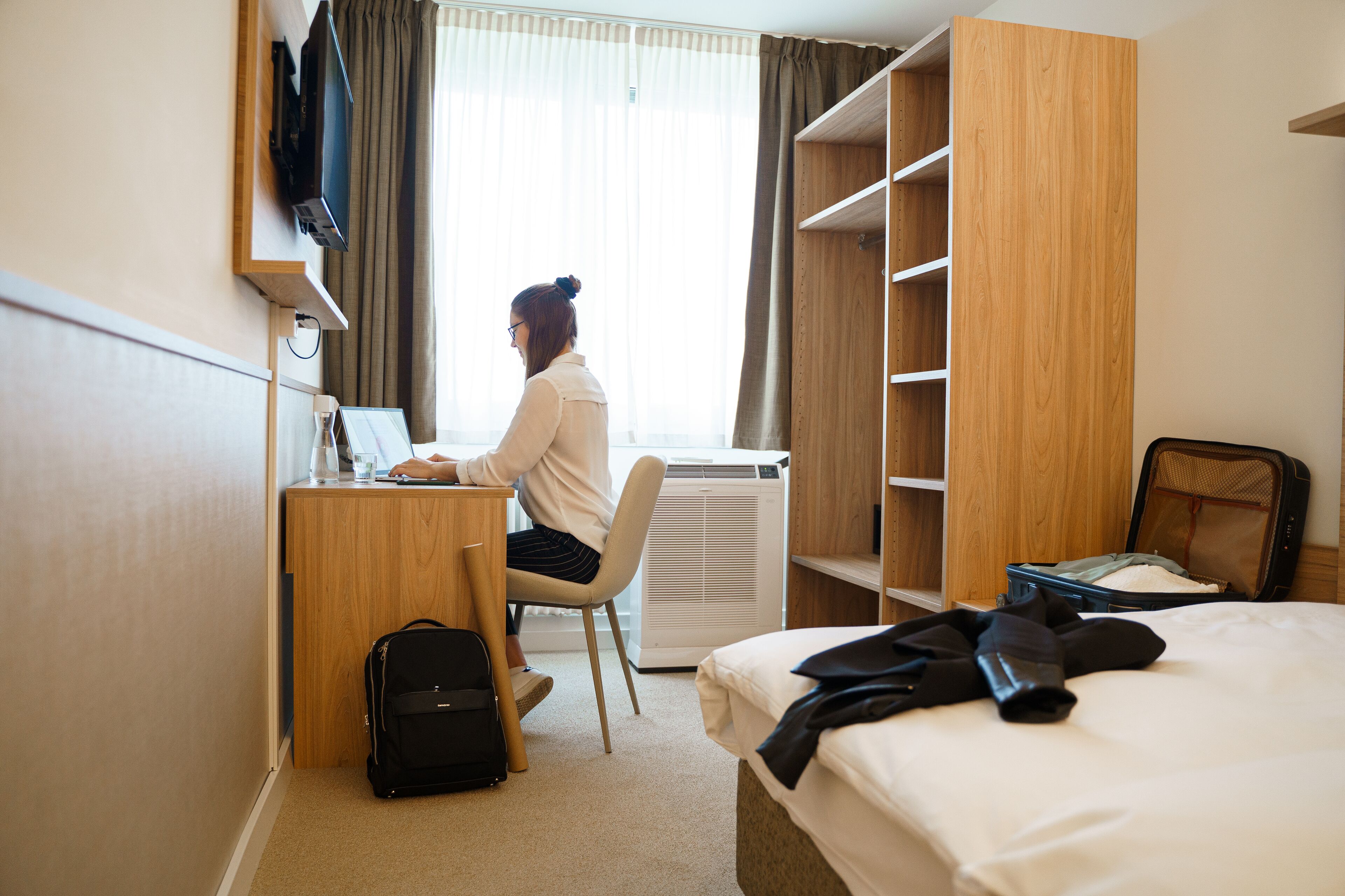 Ein Hotelzimmer mit Schreibtisch, Stuhl, Bett, Kleiderschrank und Koffer.