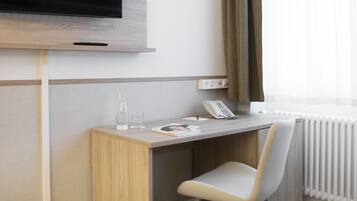 Habitación de hotel con un escritorio de madera, una silla, un televisor de pantalla plana montado en la pared, un teléfono, un vaso de agua y un radiador.