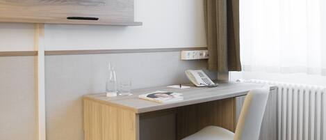 Habitación de hotel con un escritorio de madera, una silla, un televisor de pantalla plana montado en la pared, un teléfono, un vaso de agua y un radiador.