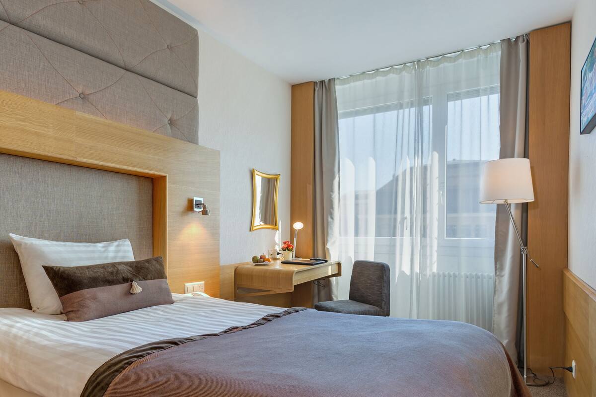 Hotellihuone, jossa on suuri sänky, työpöytä lampun kanssa, tuoli ja ikkuna, jossa on verhot.