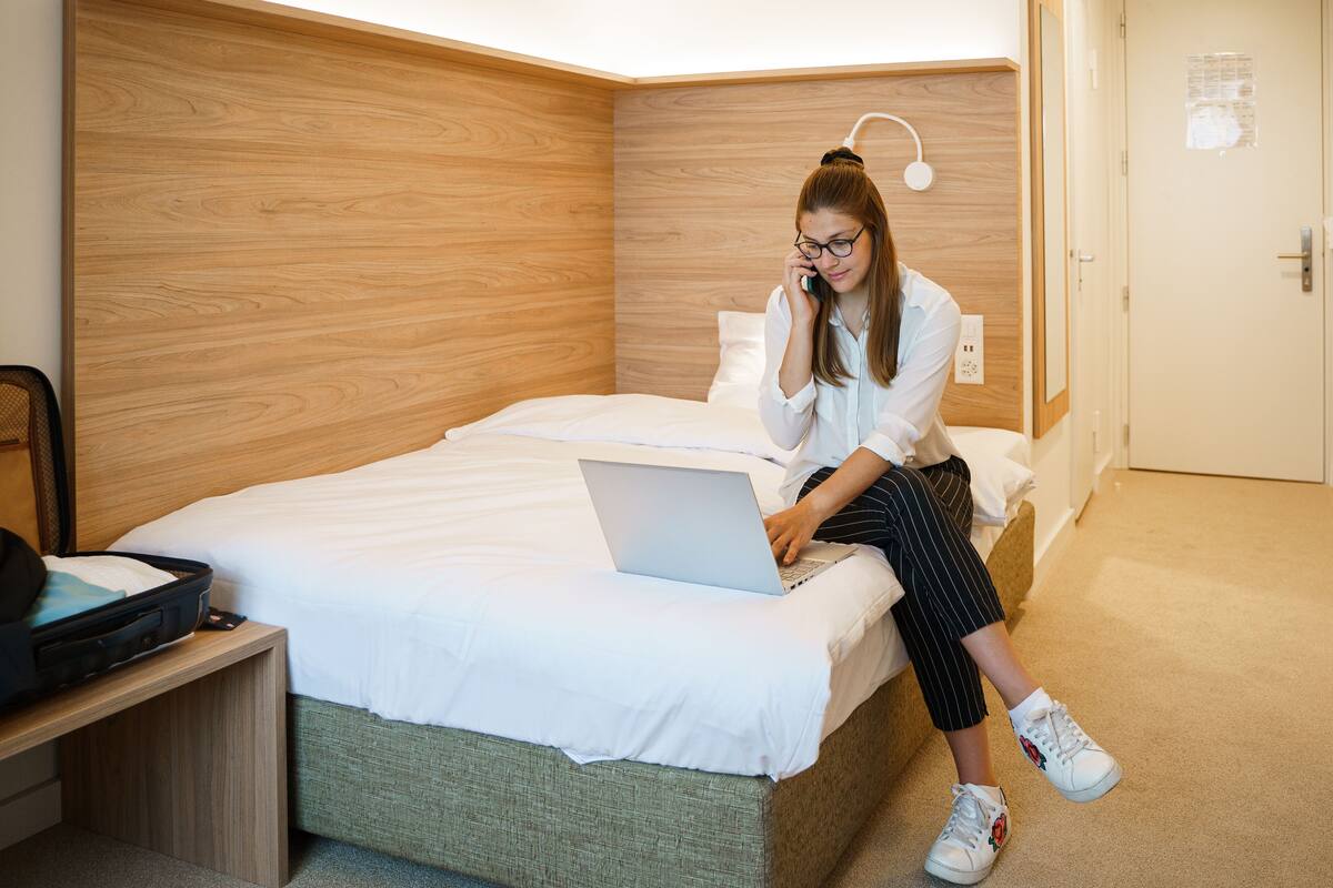 Hotellihuone, jossa on sänky, yöpöytä, jossa on matkalaukku, ja kannettava tietokone.