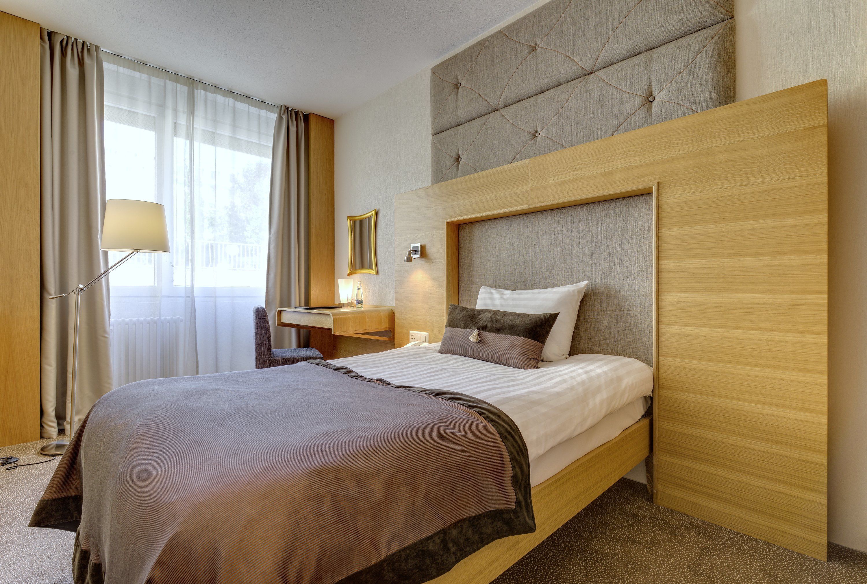 A hotel room with a bed, a desk, a chair, a lamp, and a window with curtains.