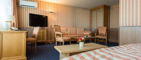 A hotel room with a large bed, a sofa, a desk with a lamp, a television, and a small table with a vase of flowers.