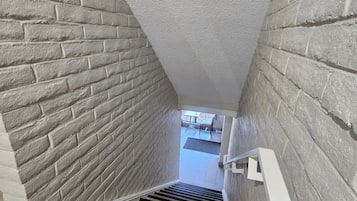 A narrow stairway with a brick wall on one side and a white textured wall on the other, leading to a room with a tiled floor.