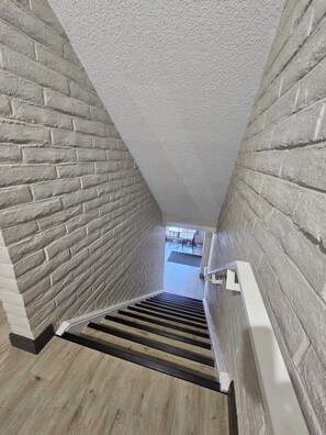 Una estrecha escalera con un muro de ladrillo a un lado y un muro blanco con textura al otro, que conduce a una habitación con suelo de baldosas.