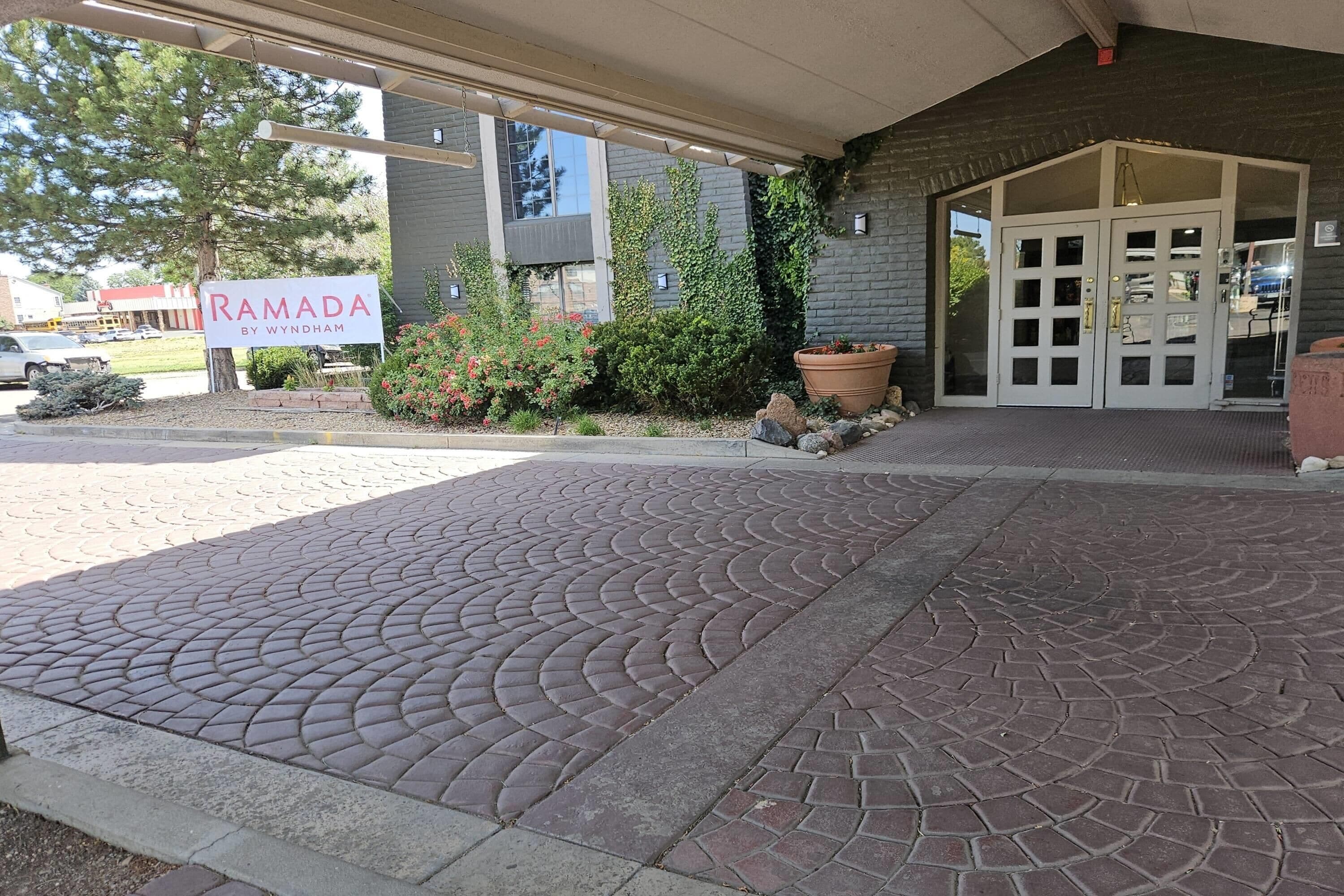 Entrada de un edificio con un sendero pavimentado, un letrero que dice 'RAMADA BY WYNDHAM' y una jardinera grande con plantas.