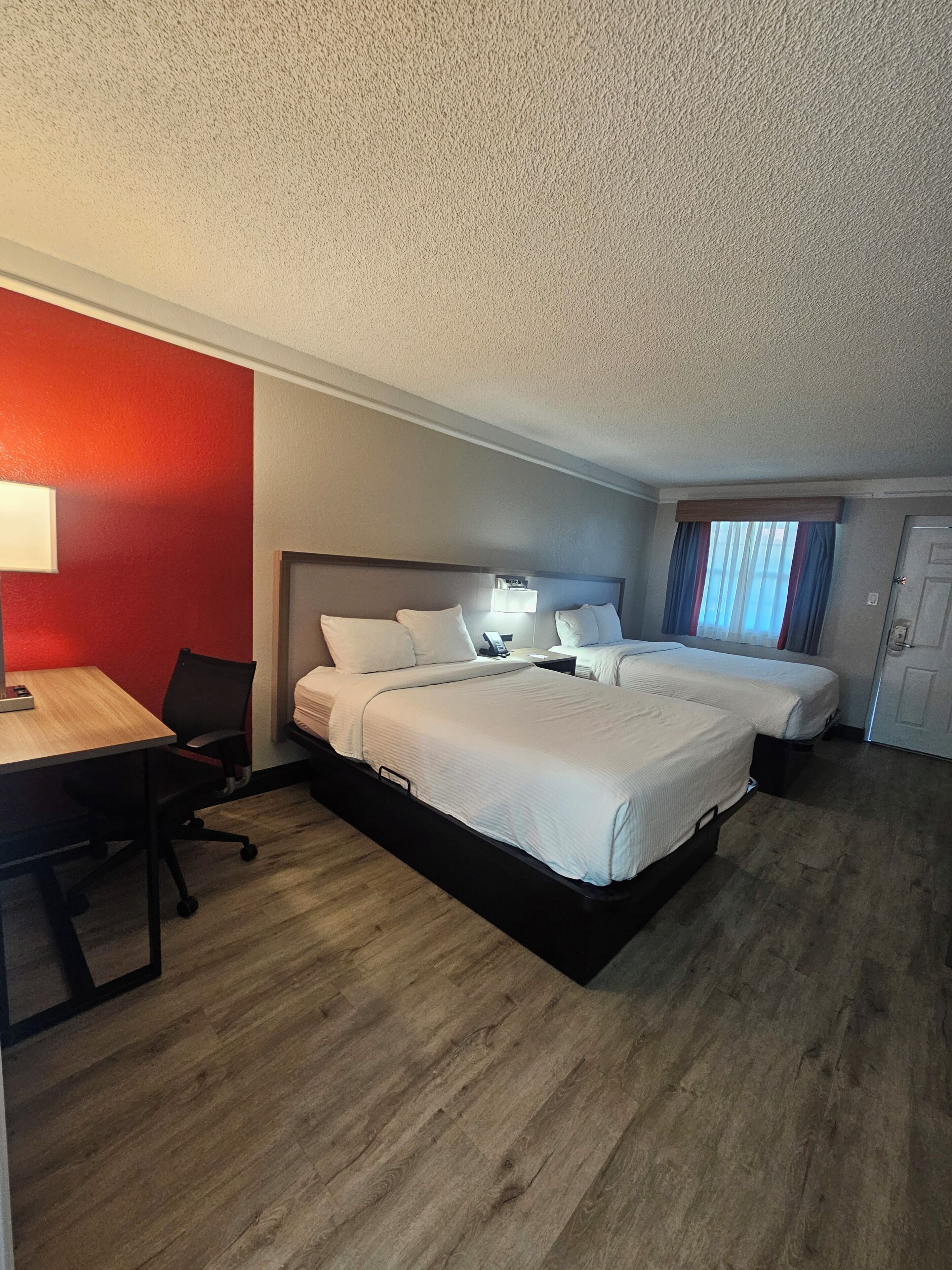Quarto de hotel com cama, mesa de cabeceira, luminária e escrivaninha com cadeira.