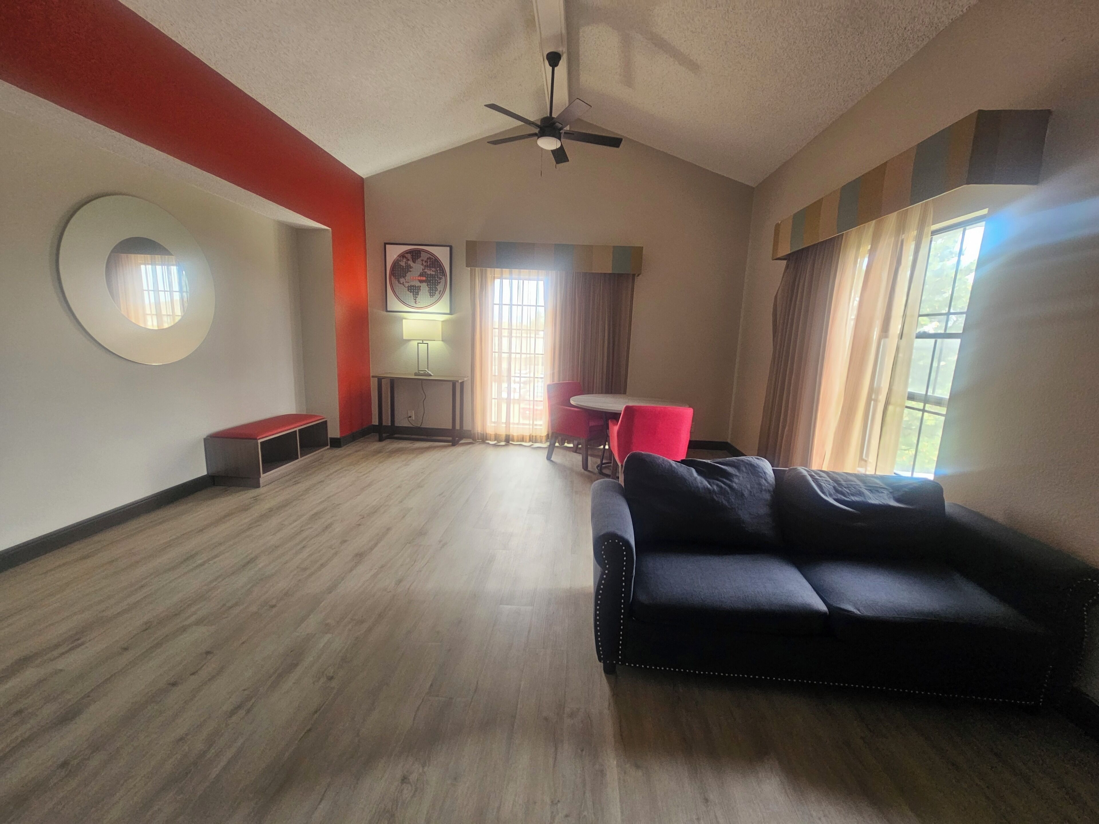 A spacious living area with a black sofa, a dining table with red chairs, a ceiling fan, and a large window with curtains.