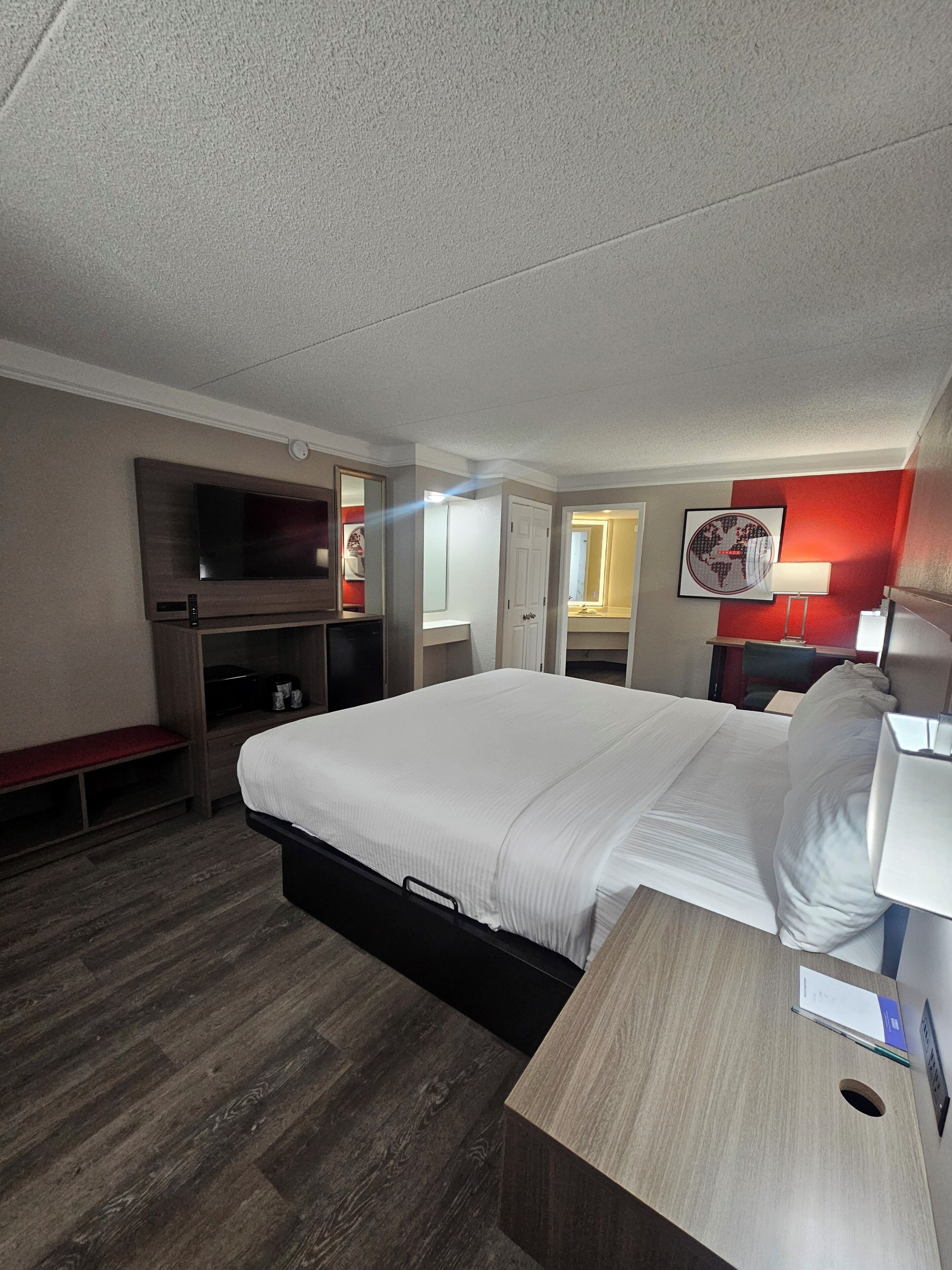 A hotel room with a bed, a desk, a TV, a mirror, and a red accent wall.